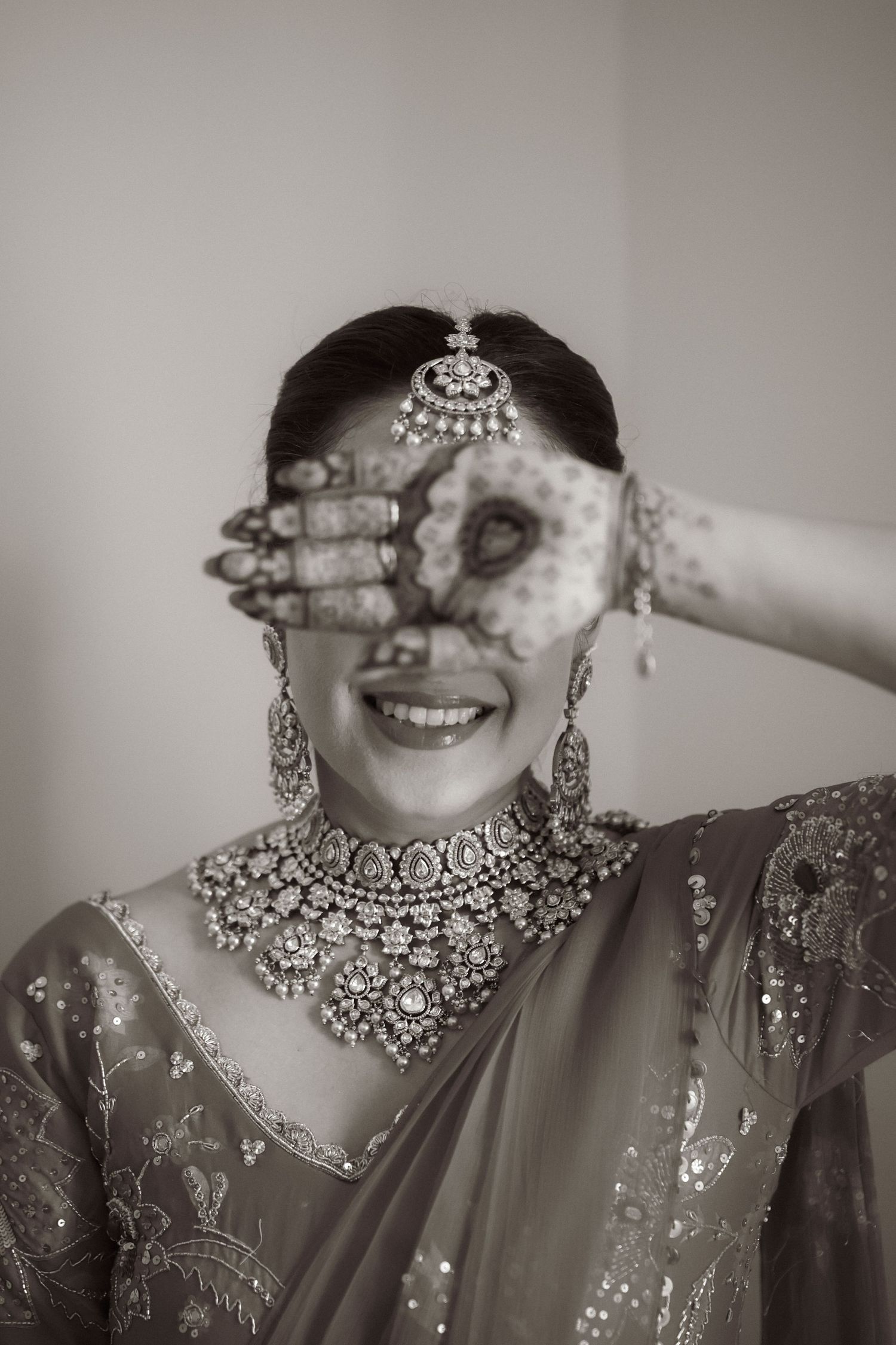 Black and white photo of smiling Indian bride in saree with mehndi hand, maang tikka, chandelier earrings, and diamond necklace.