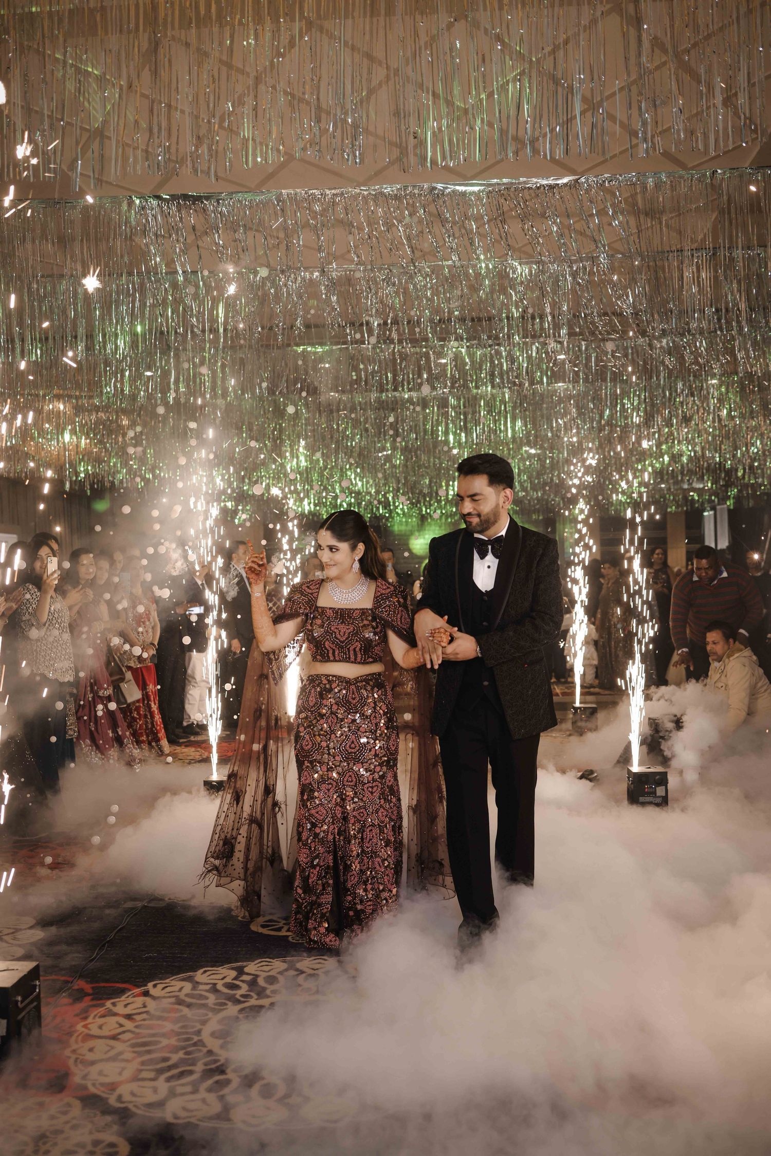 Couple makes grand entrance at wedding reception with sparklers, fog machine, and glittering silver fringe backdrop.