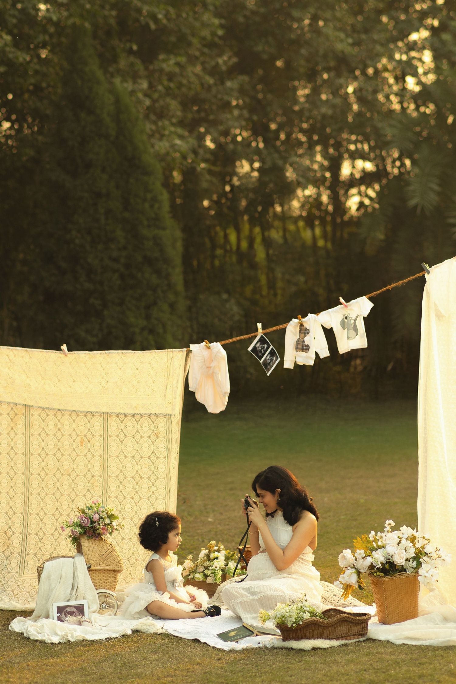 Mother and daughter in white dresses enjoy a vintage outdoor picnic photoshoot with clothesline and flowers.