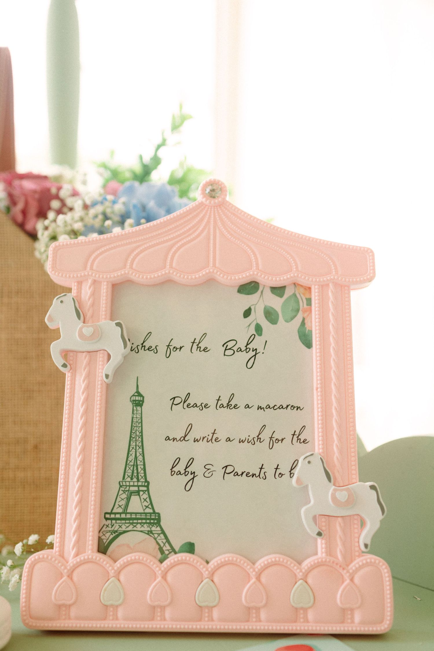 Pink carousel-themed picture frame displaying a baby shower wish card with Eiffel Tower design on a decorated table.