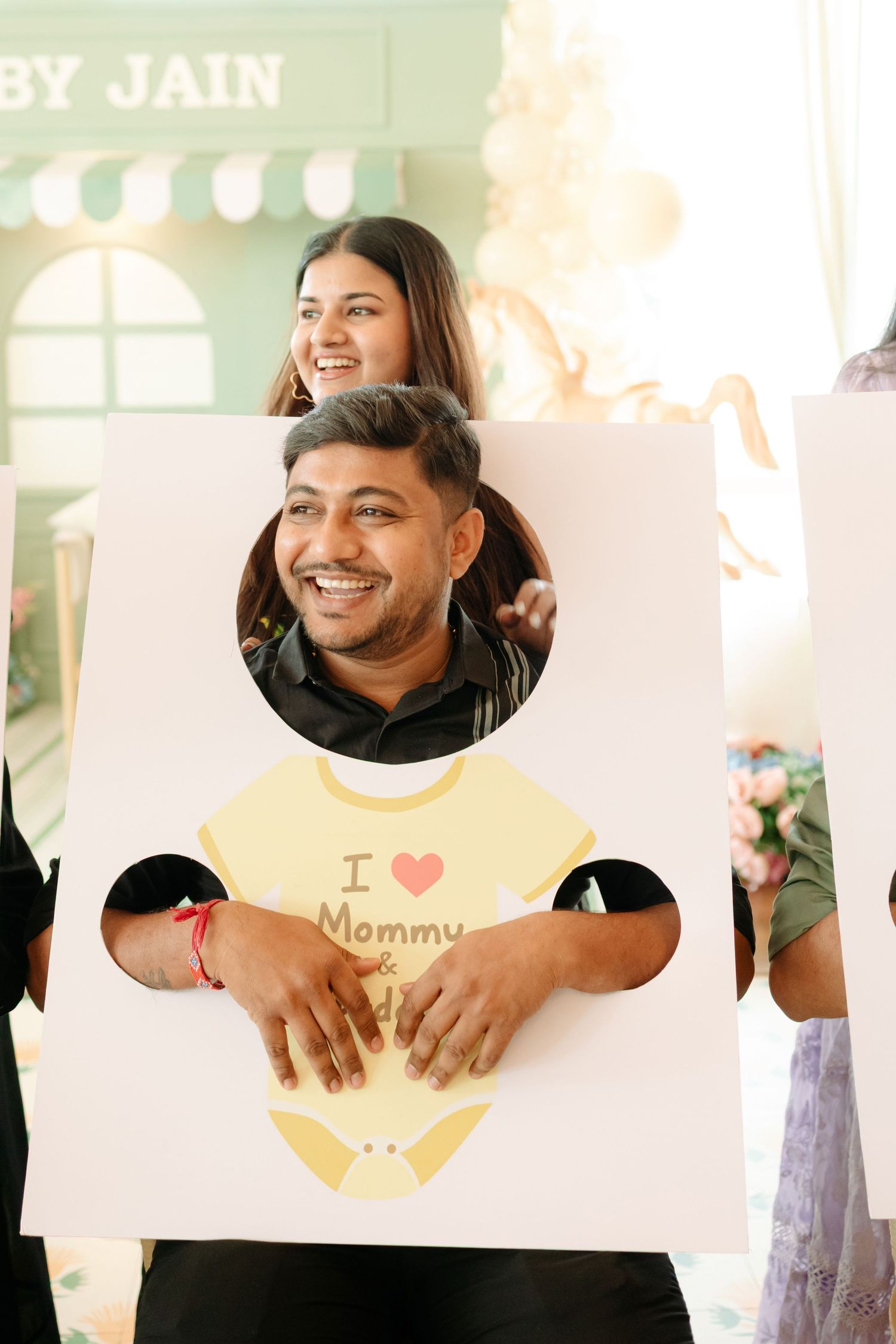 Man smiling through a baby onesie cutout board at a baby shower event with guests behind him.