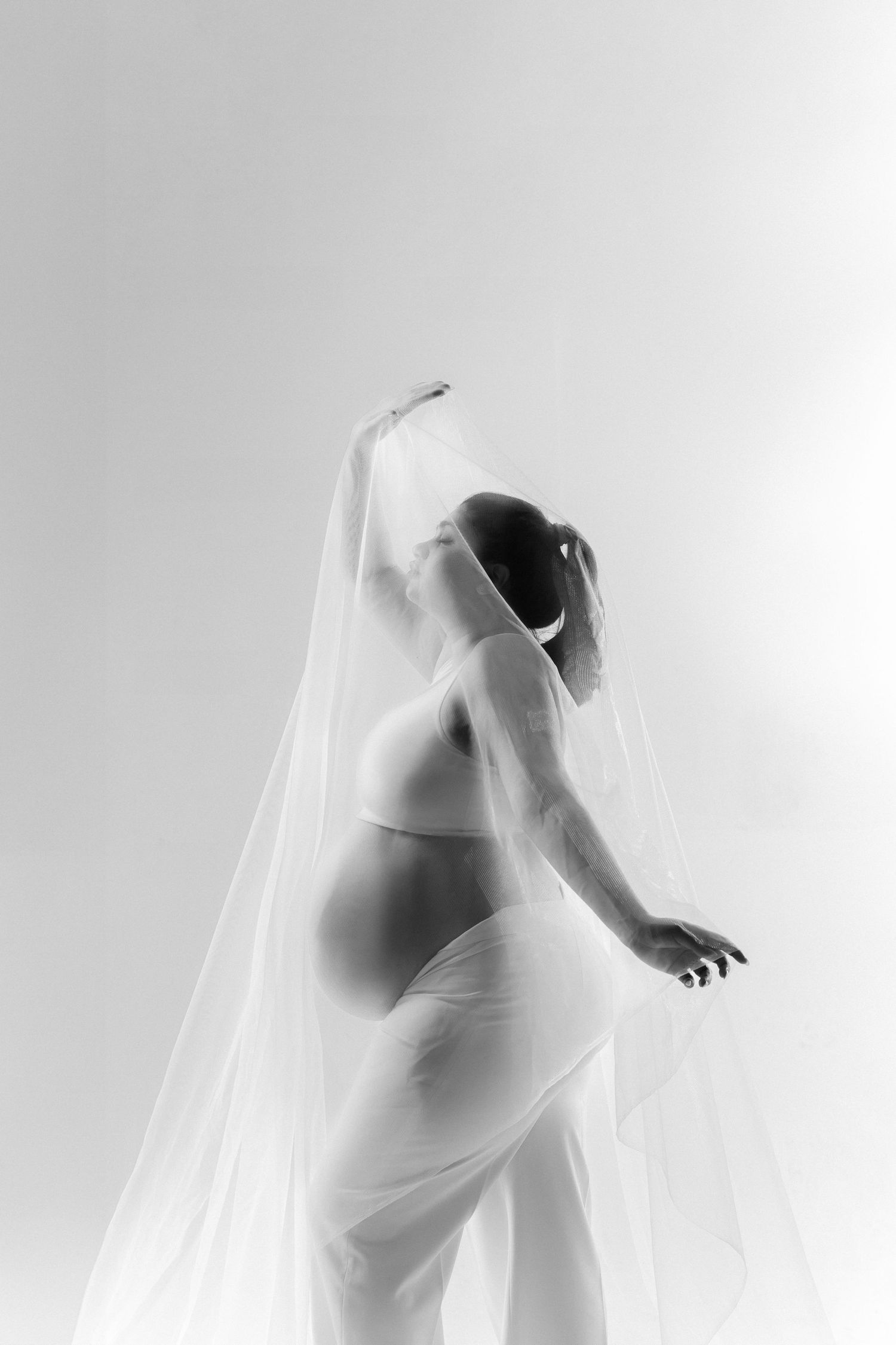 Black and white photo of a pregnant woman in lingerie draped with sheer fabric, posing gracefully against a light background.