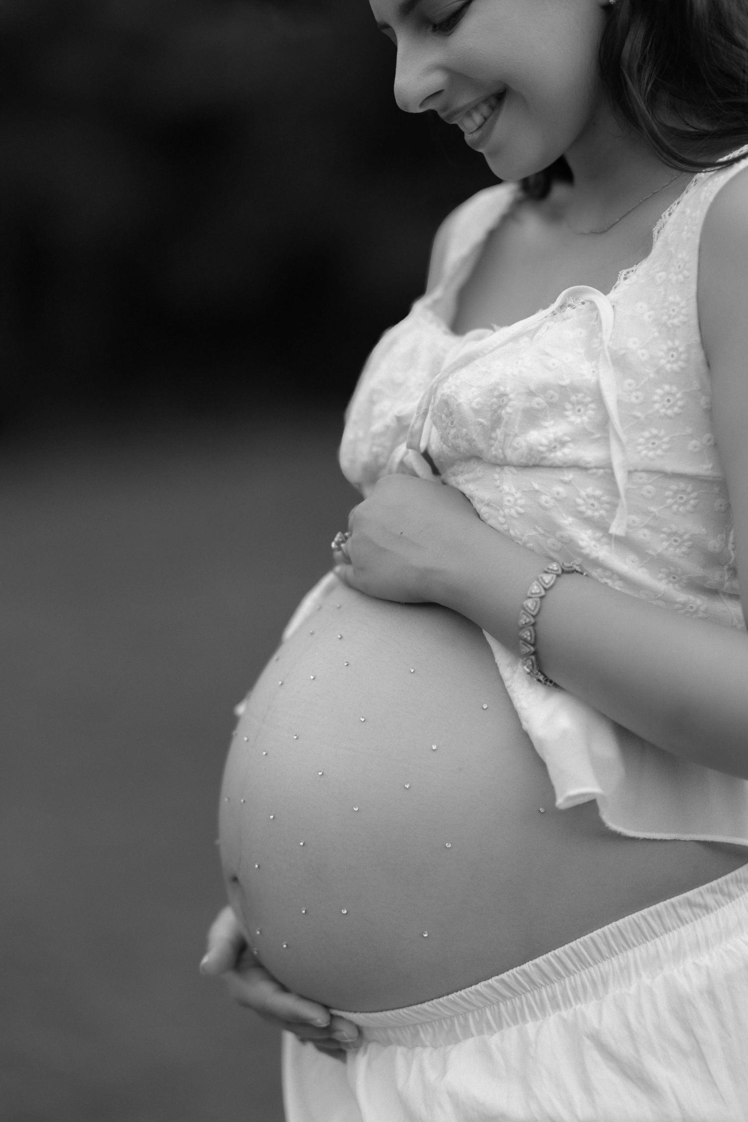 Black and white maternity photo series showing a pregnant belly in a white lace dress with delicate details.