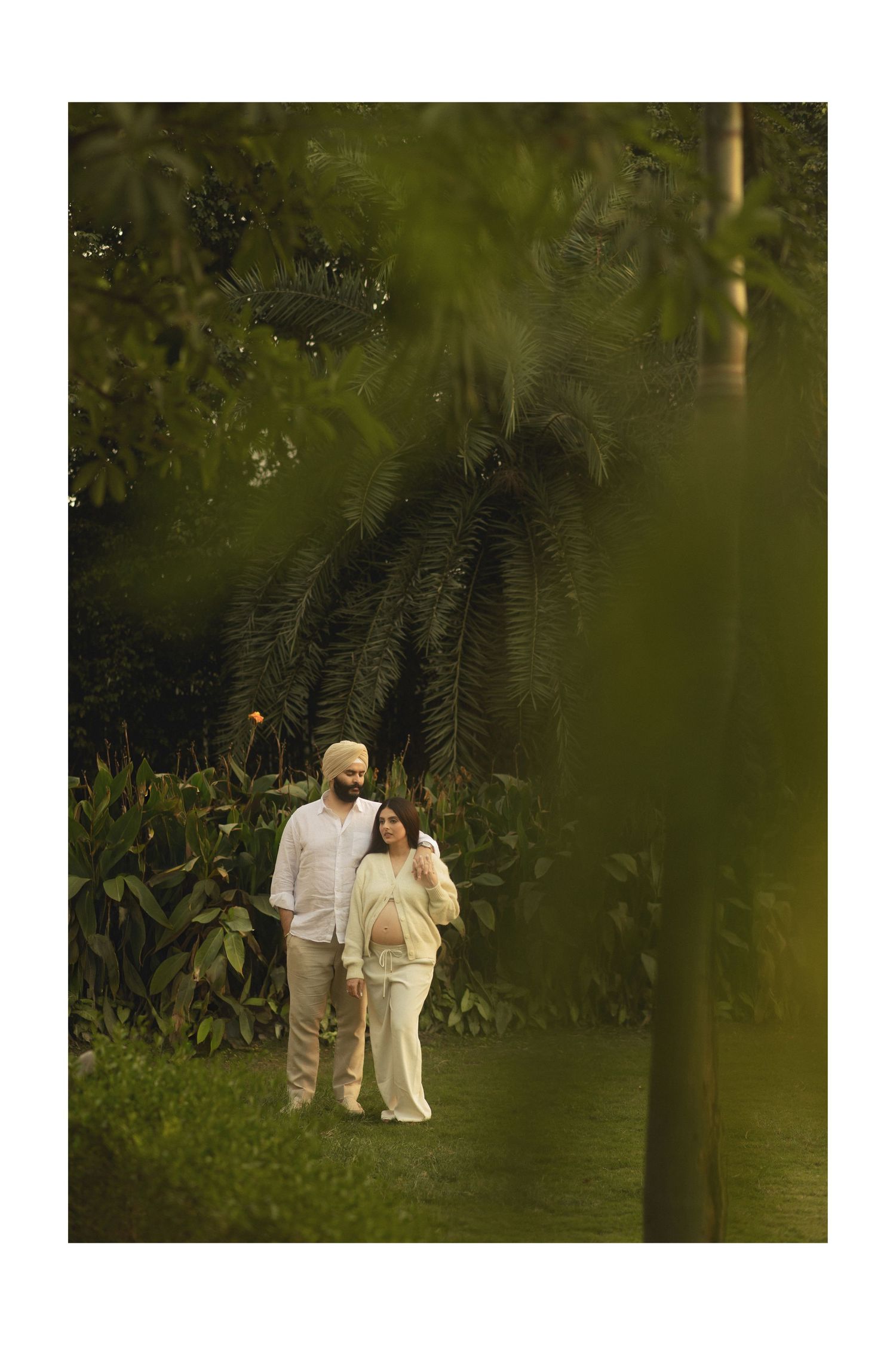 A couple in white attire walks together through a lush garden setting during a romantic outdoor photoshoot.