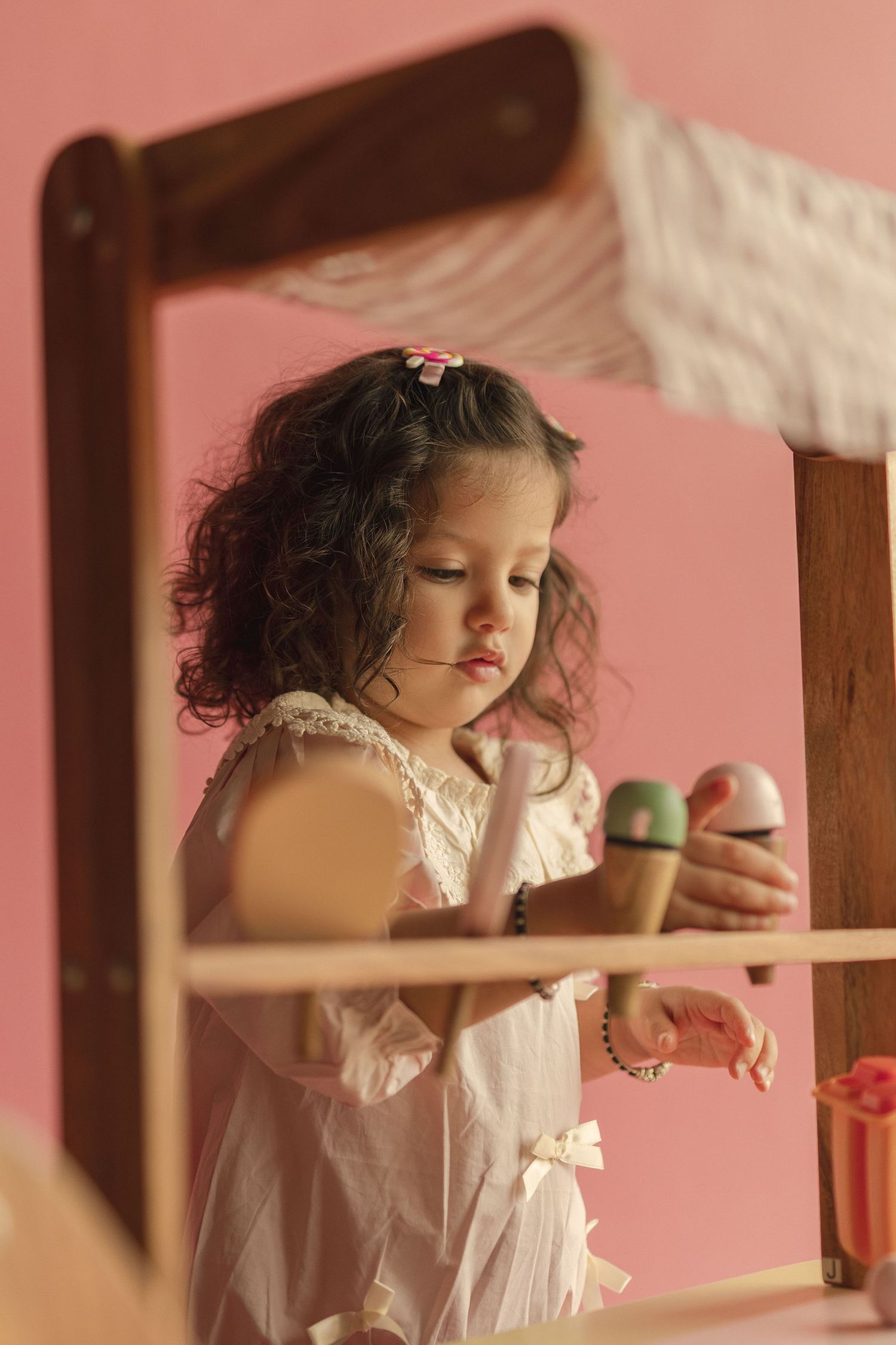 Young girl with curly hair in white dress playing with colorful toy ice cream cones against pink background.