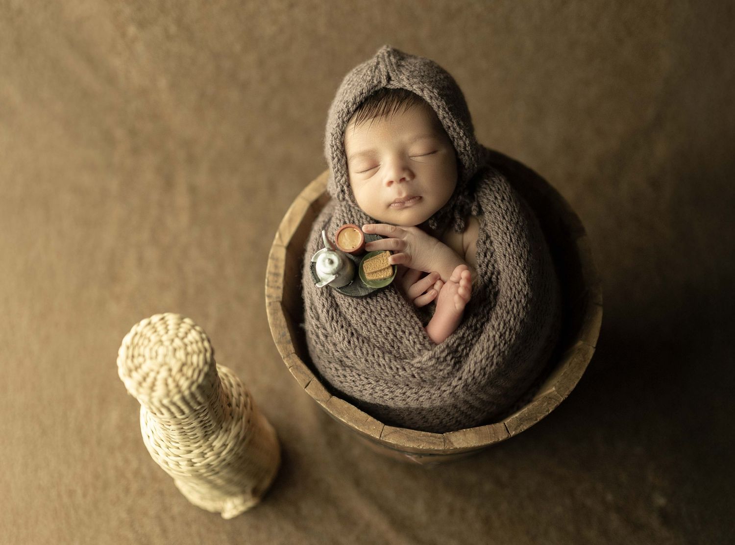 Sleeping newborn baby wrapped in gray knit blanket inside wooden bowl with owl figurine beside.