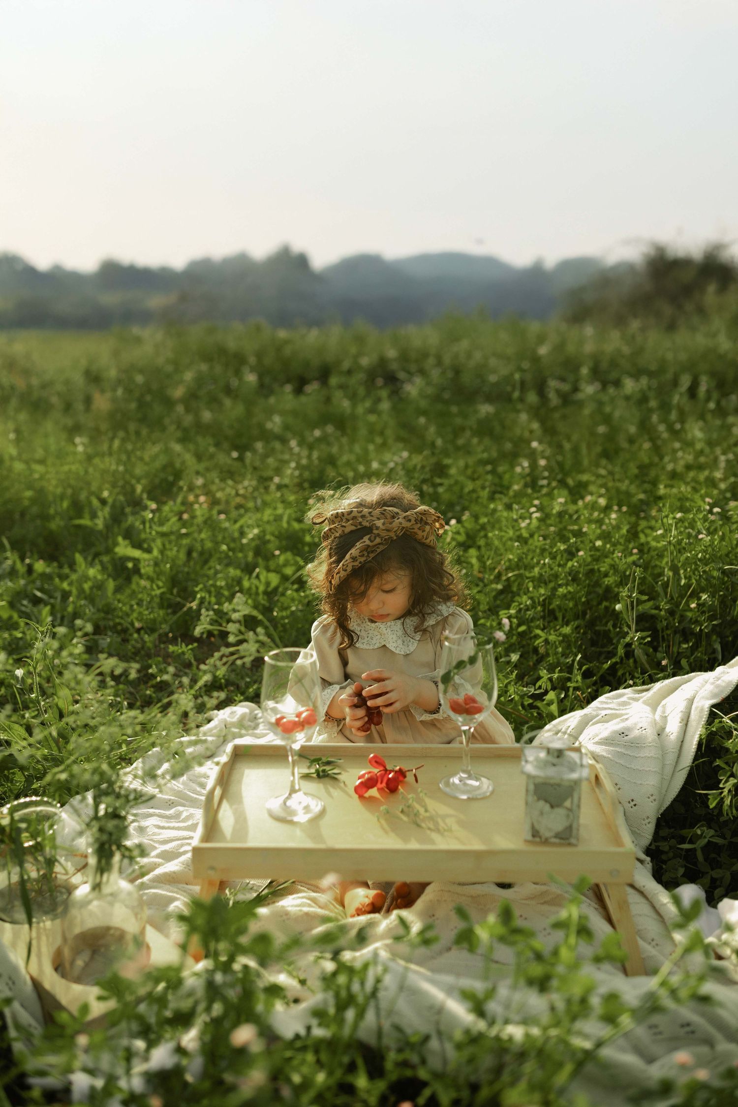A young girl with a floral crown sits on a blanket in a green field, arranging flowers on a wooden tray.
