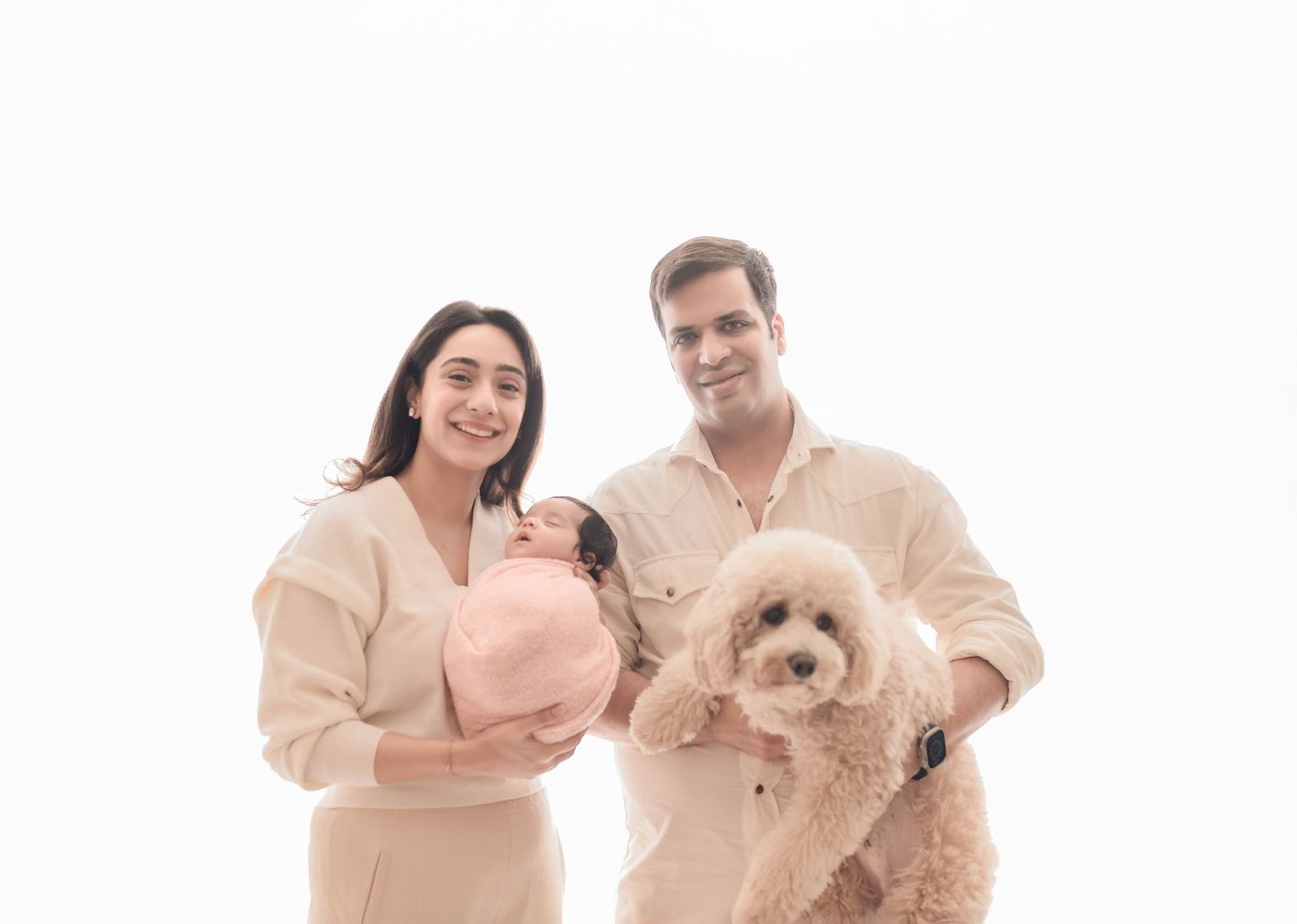 A couple poses with a newborn baby and a fluffy beige poodle against a white background.