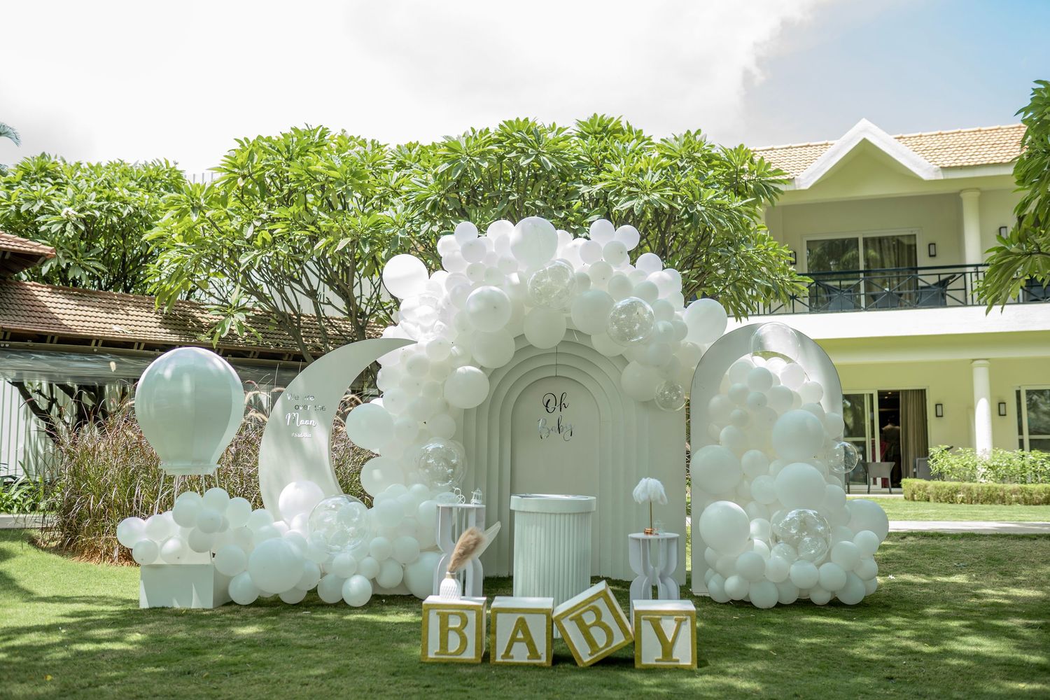 Elegant baby shower setup with white balloon arch, floral decor, and BABY letter blocks on green lawn.