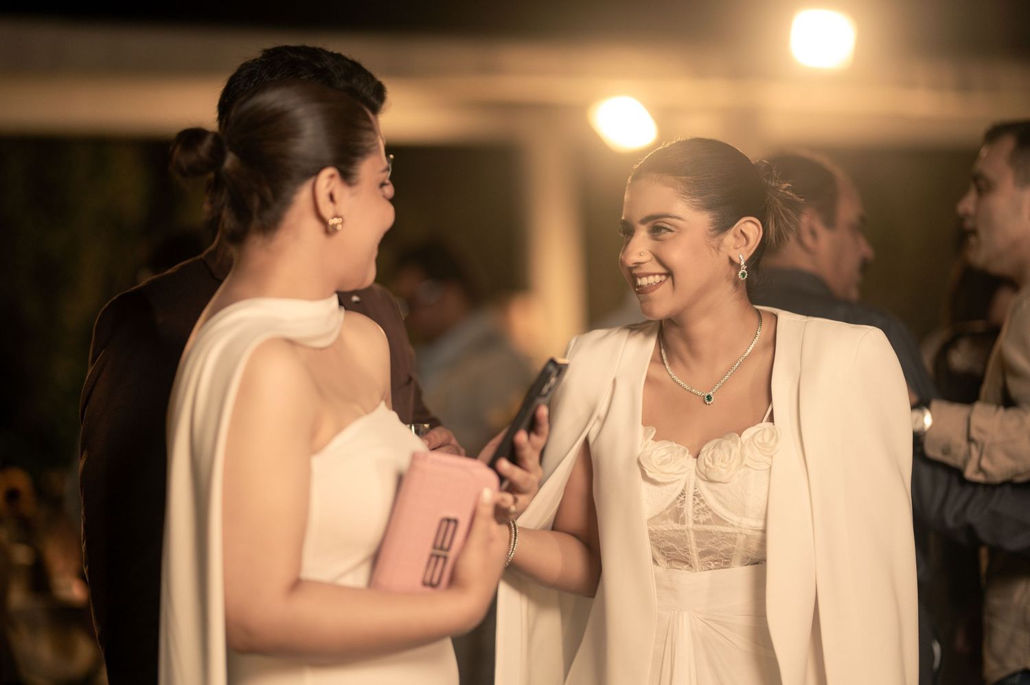 Two women in elegant white outfits smile and chat at an upscale evening event.