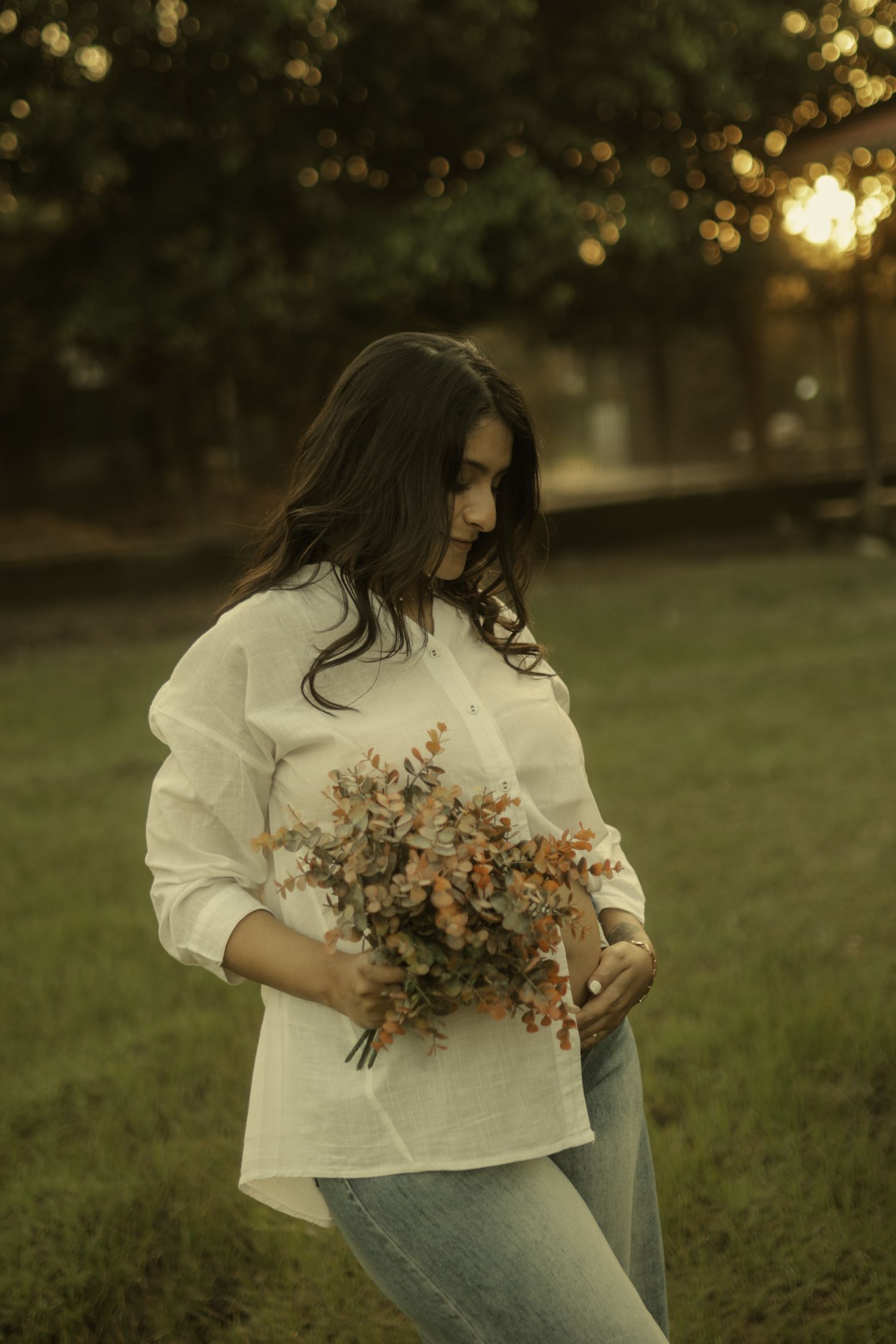 Woman in white linen shirt holding wildflower bouquet standing on green grass at golden hour sunset.