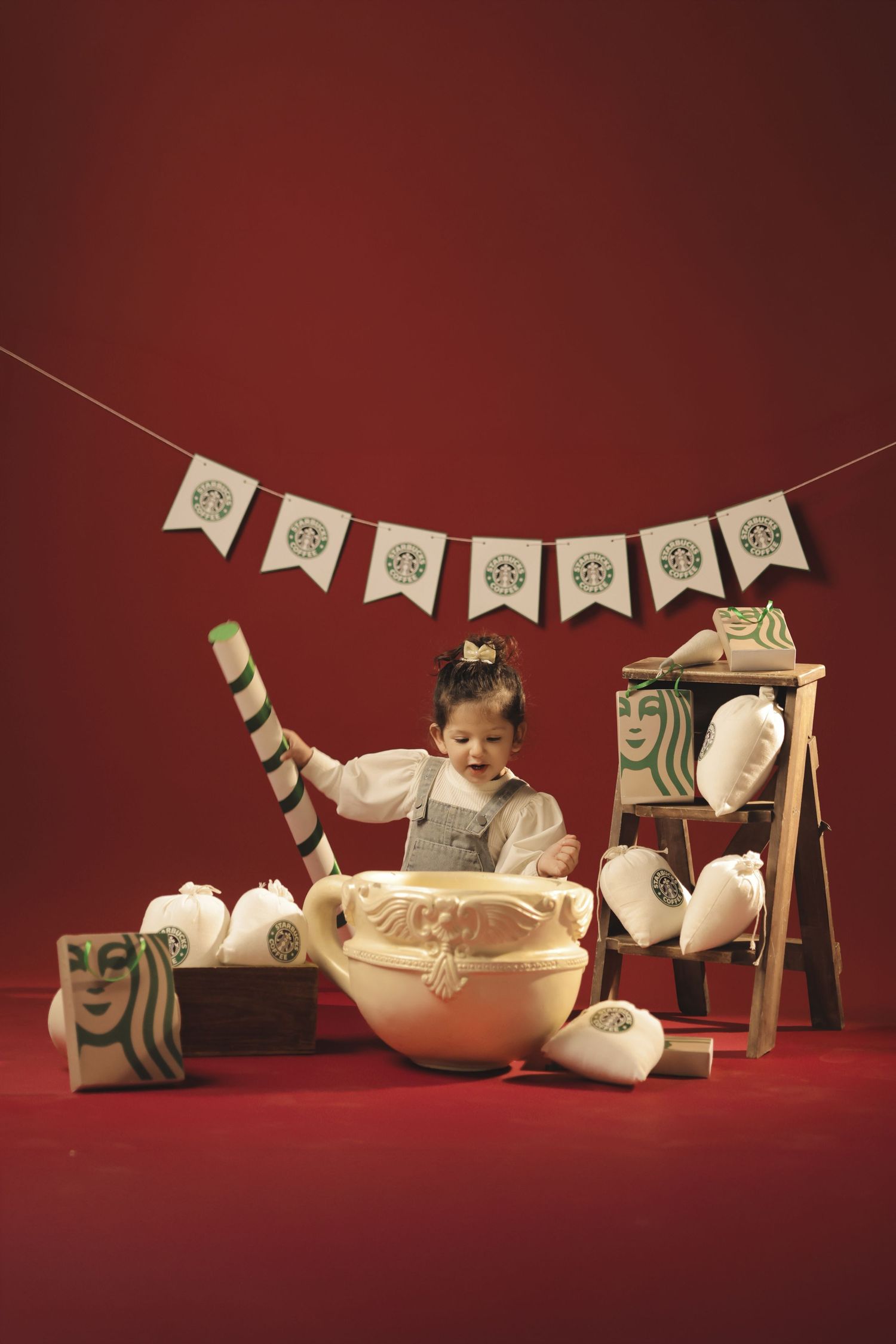 Toddler girl surrounded by Starbucks-themed props, pillows, and bunting in a festive red studio photo shoot.