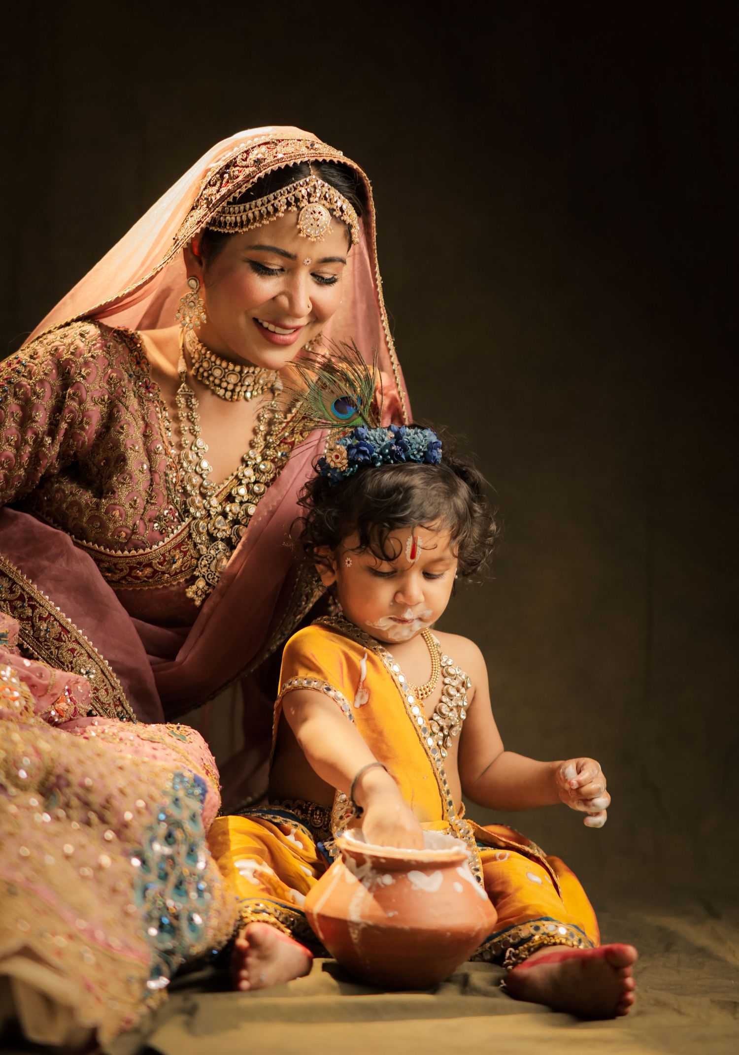 Woman in traditional Indian bridal attire smiles at a young child dressed as Krishna playing with a clay pot.