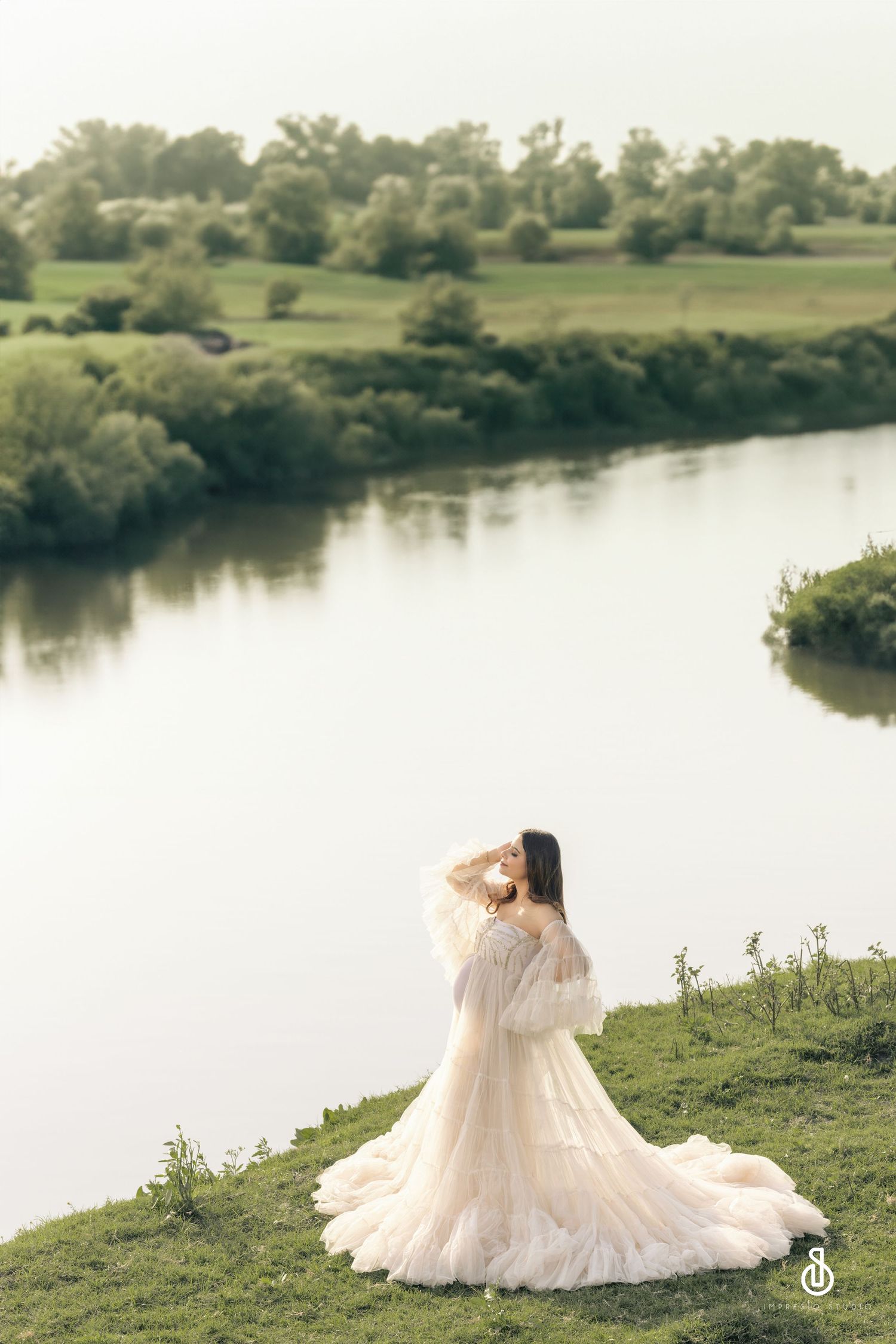 A person in a maternity gown stands on a grassy hill overlooking a serene river on a misty day.