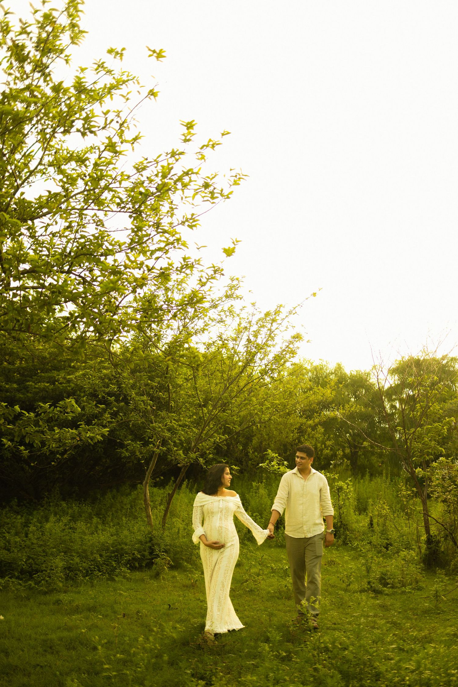 A couple walks hand in hand through a sunlit garden wearing elegant white clothing with trees creating a romantic backdrop.