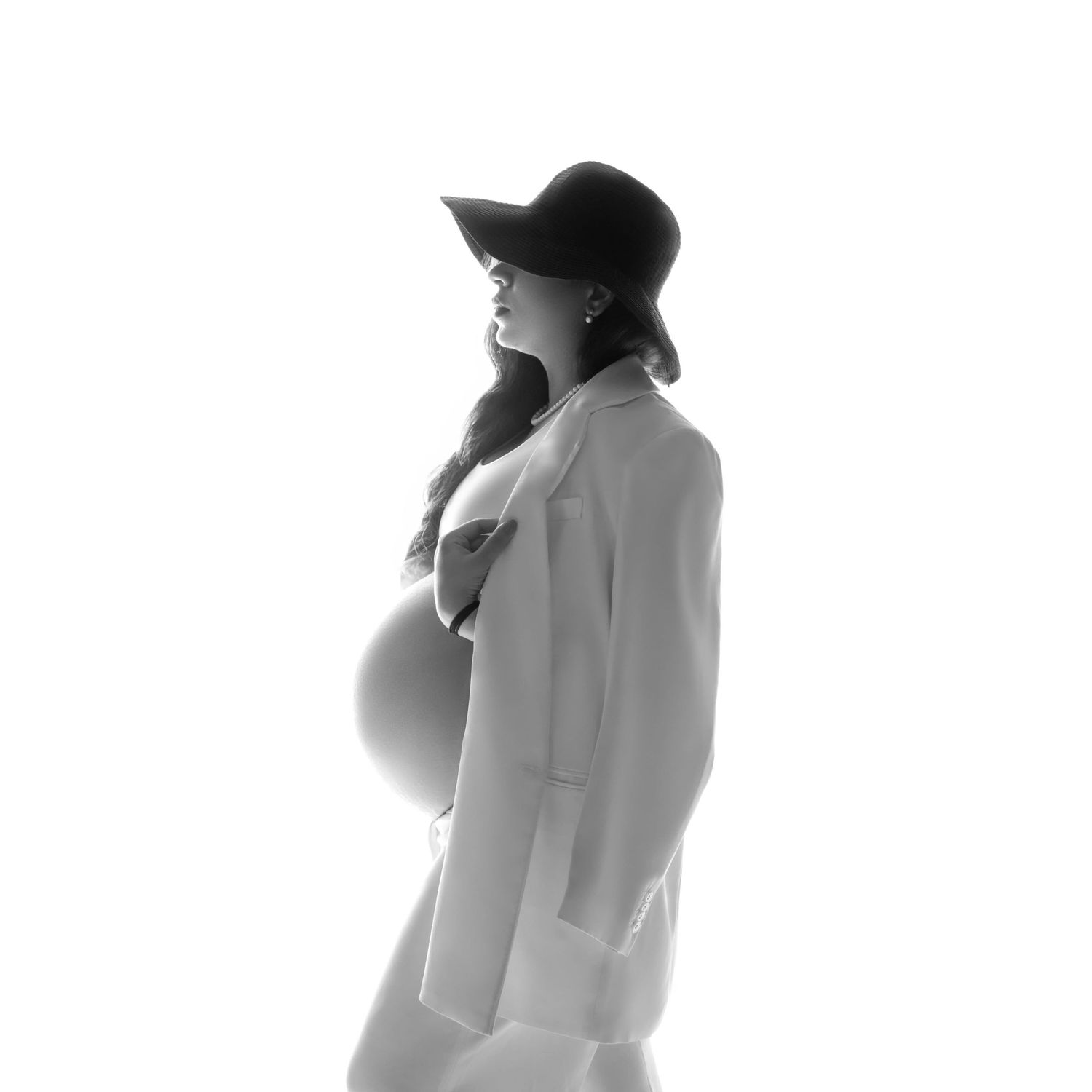 Black and white artistic maternity photograph of a woman in a light colored coat and dark hat looking off to the side.