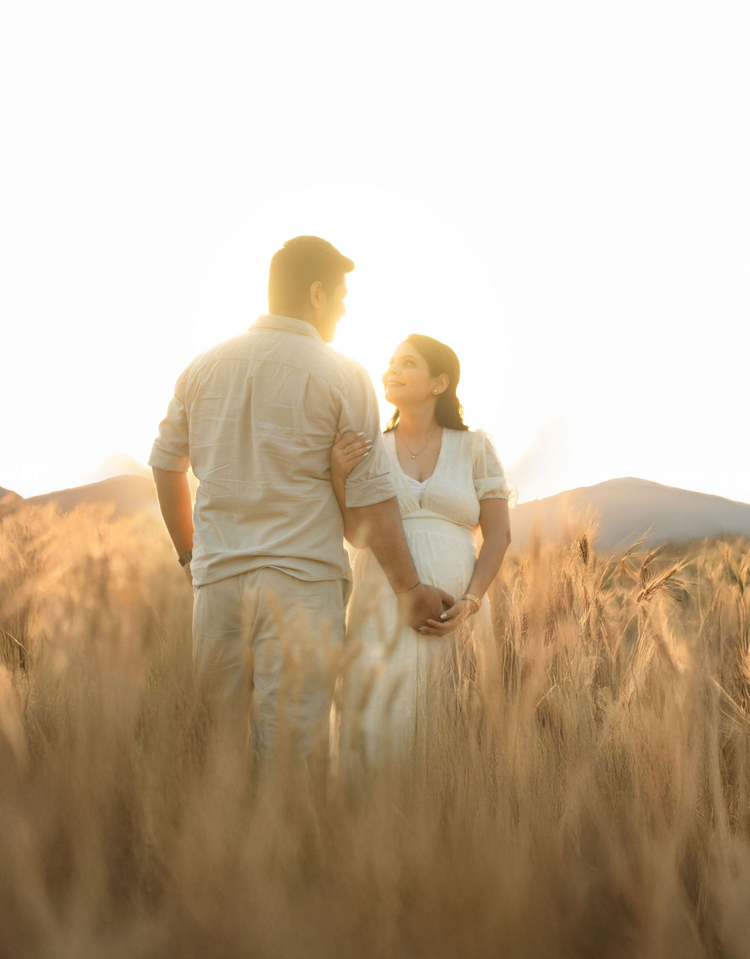 Couple standing together in golden wheat field at sunset with mountains in background during maternity photoshoot.