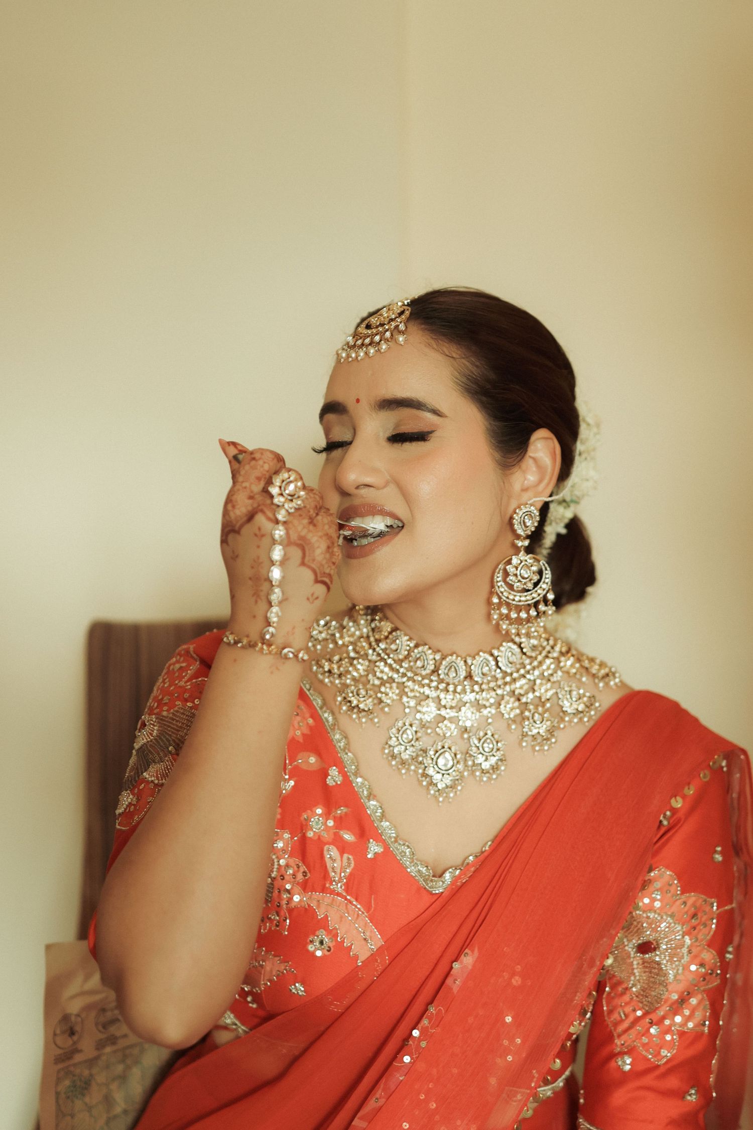 Bride in red saree wearing kundan jewelry, maang tikka, and earrings, smiling gracefully during bridal preparation.