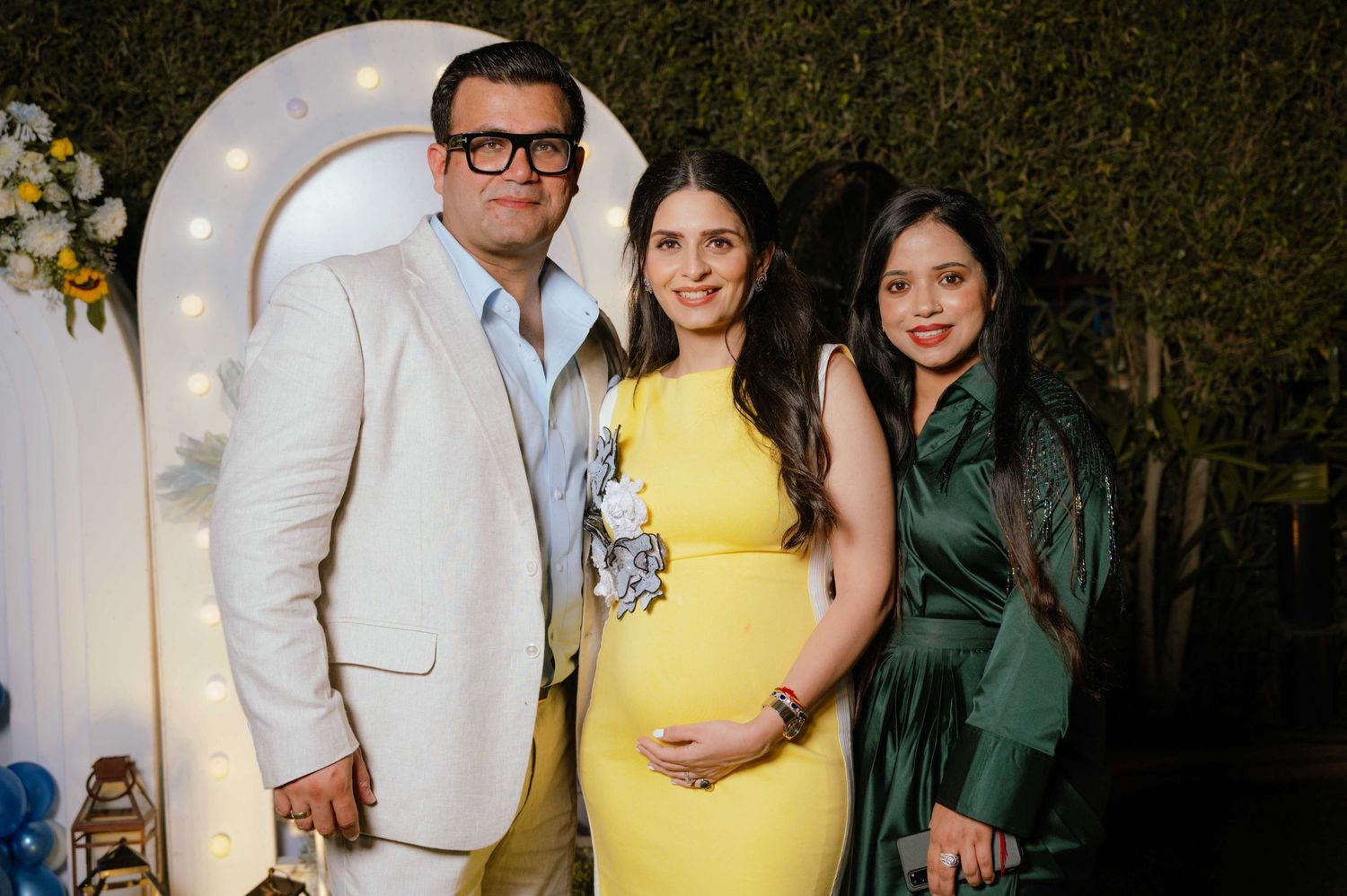 Pregnant woman in yellow dress poses with man in white suit and woman in green dress at baby shower.