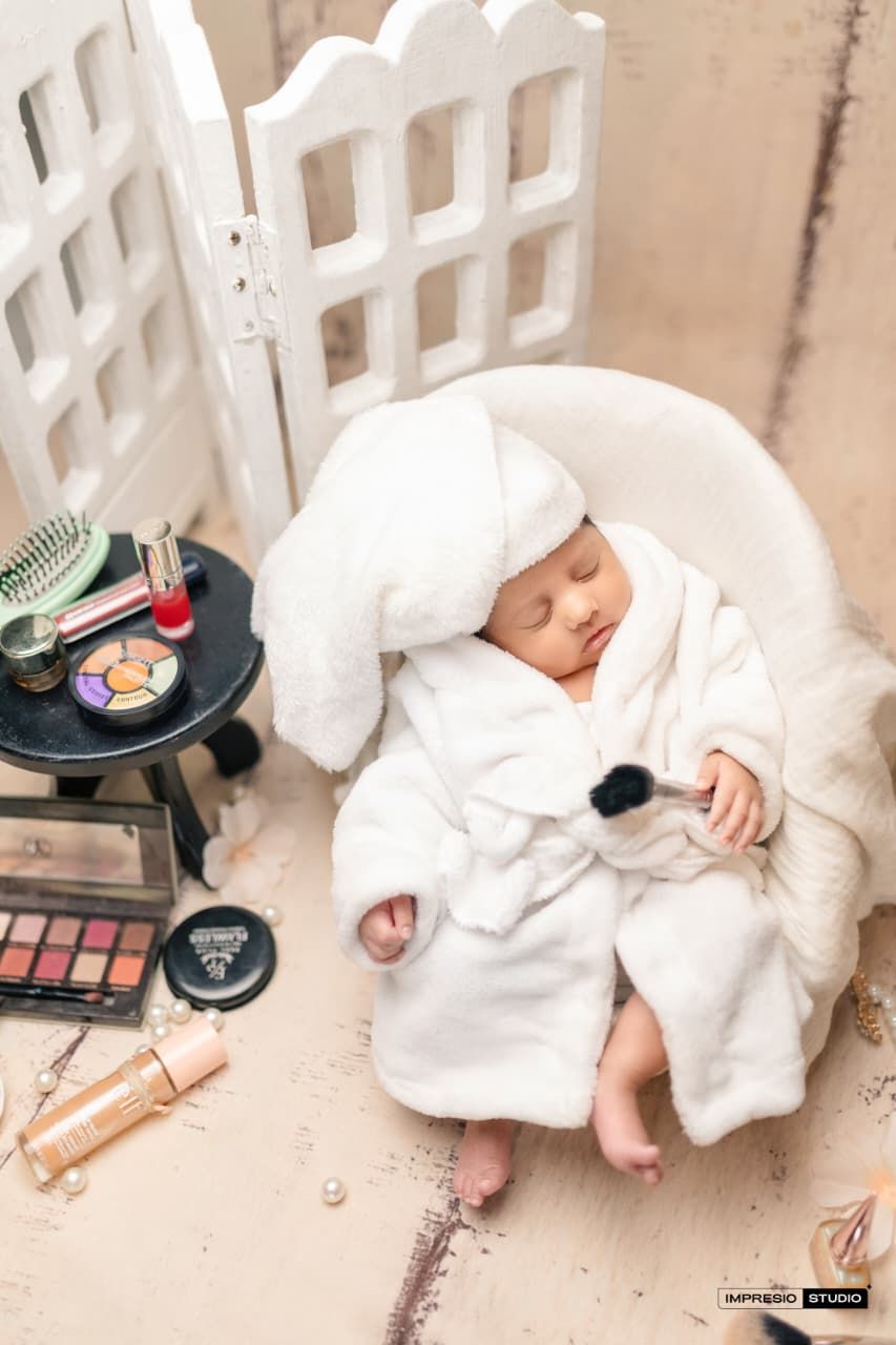 Newborn baby in white fluffy robe and towel turban sleeping in mini chair surrounded by makeup products.