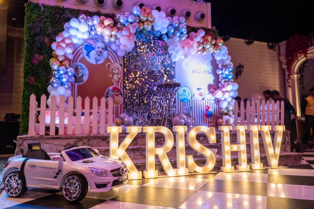 Colorful balloon arch decorates a birthday party setup with illuminated KRISHIV letters and a toy car on the dance floor.