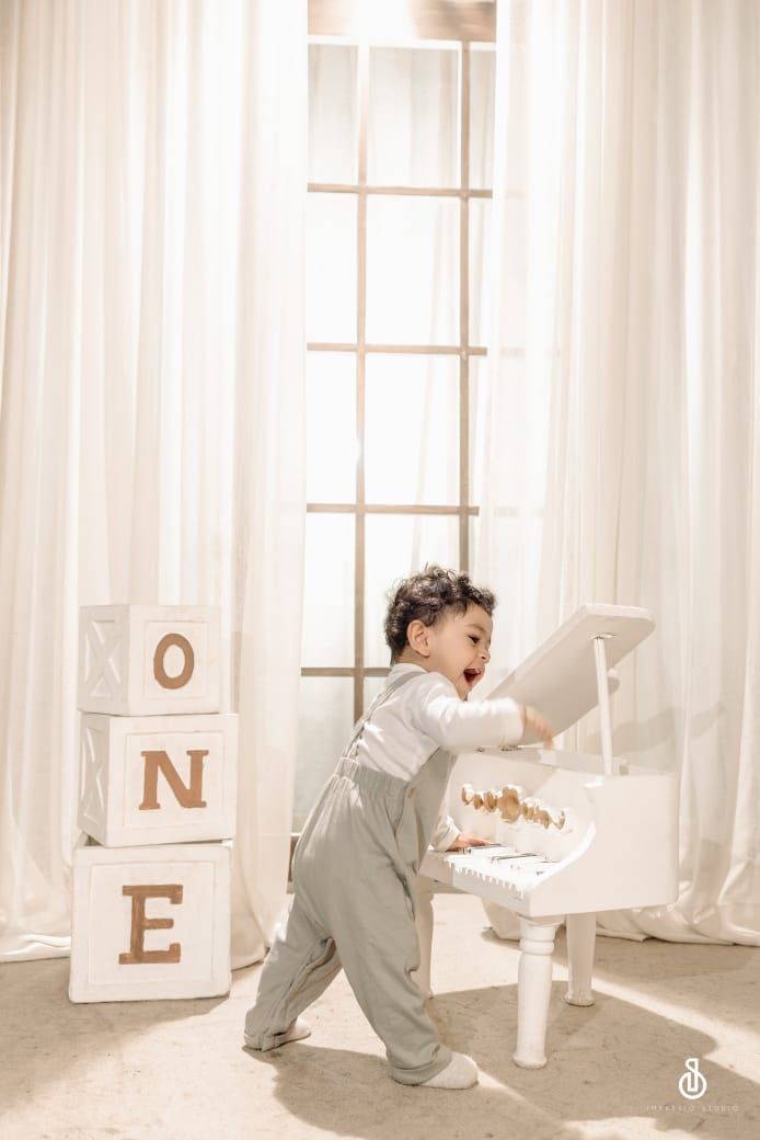 Toddler boy in gray overalls playing with a white toy grand piano next to letter blocks spelling ONE for first birthday.