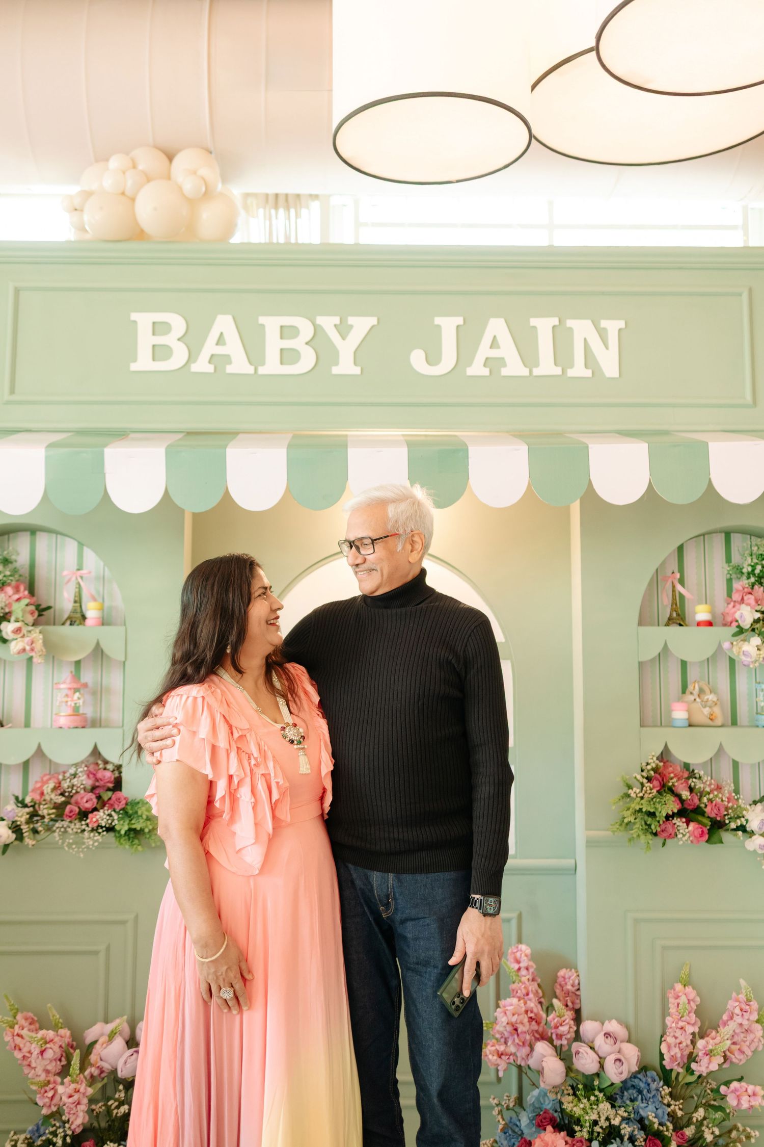 Couple smiling in front of a mint green Baby Jain baby shower backdrop with floral arrangements.