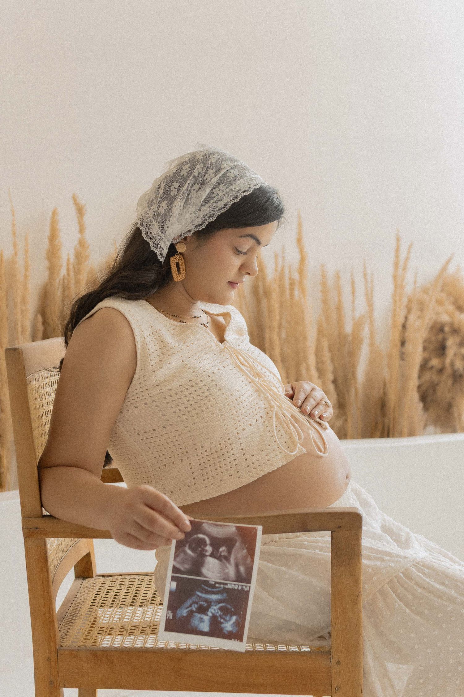 Pregnant woman in cream crochet dress sits in wooden chair, tenderly holding ultrasound photos while touching her baby bump.