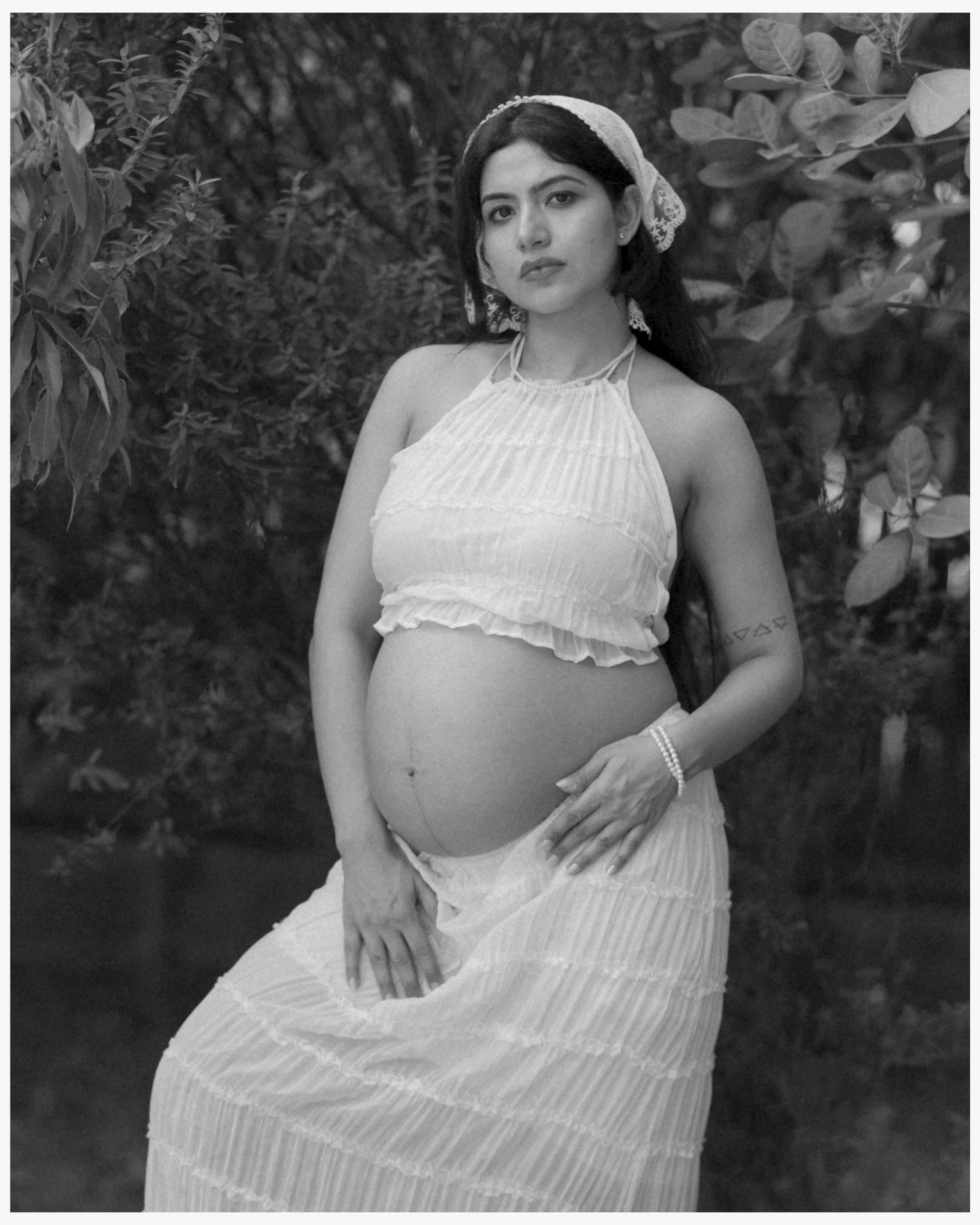 Black and white maternity photo of a pregnant woman in white halter crop top and skirt outdoors.