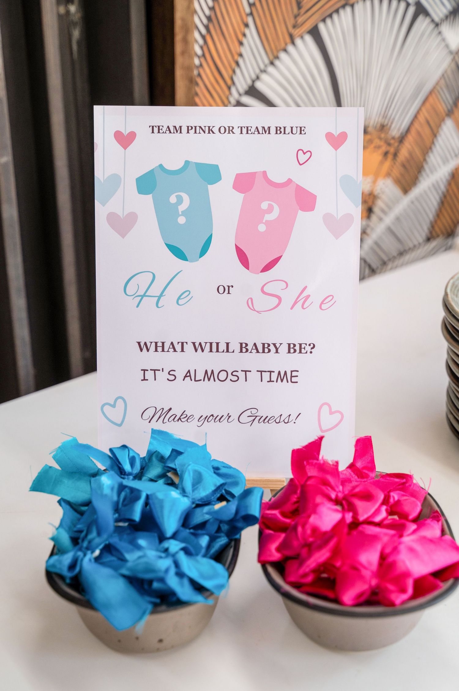 Gender reveal party display with blue and pink tissue paper bowls beside a He or She baby gender guess sign.