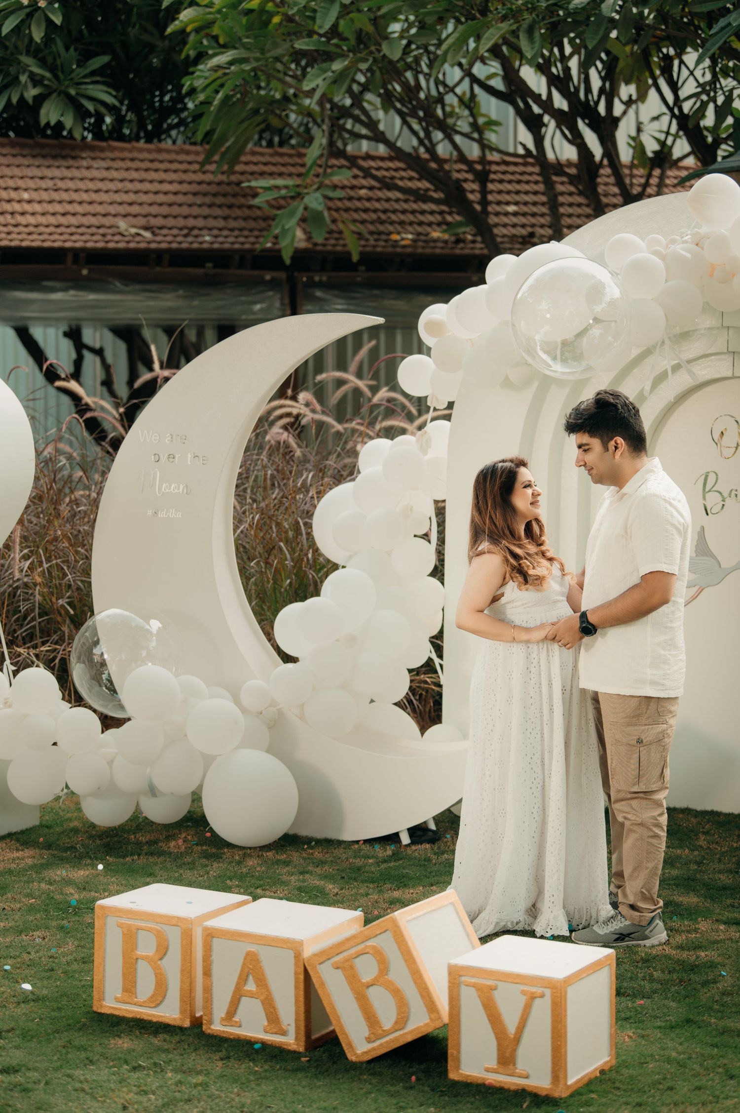 Couple at outdoor baby shower with white balloon moon backdrop and large wooden BABY letter blocks on green lawn.