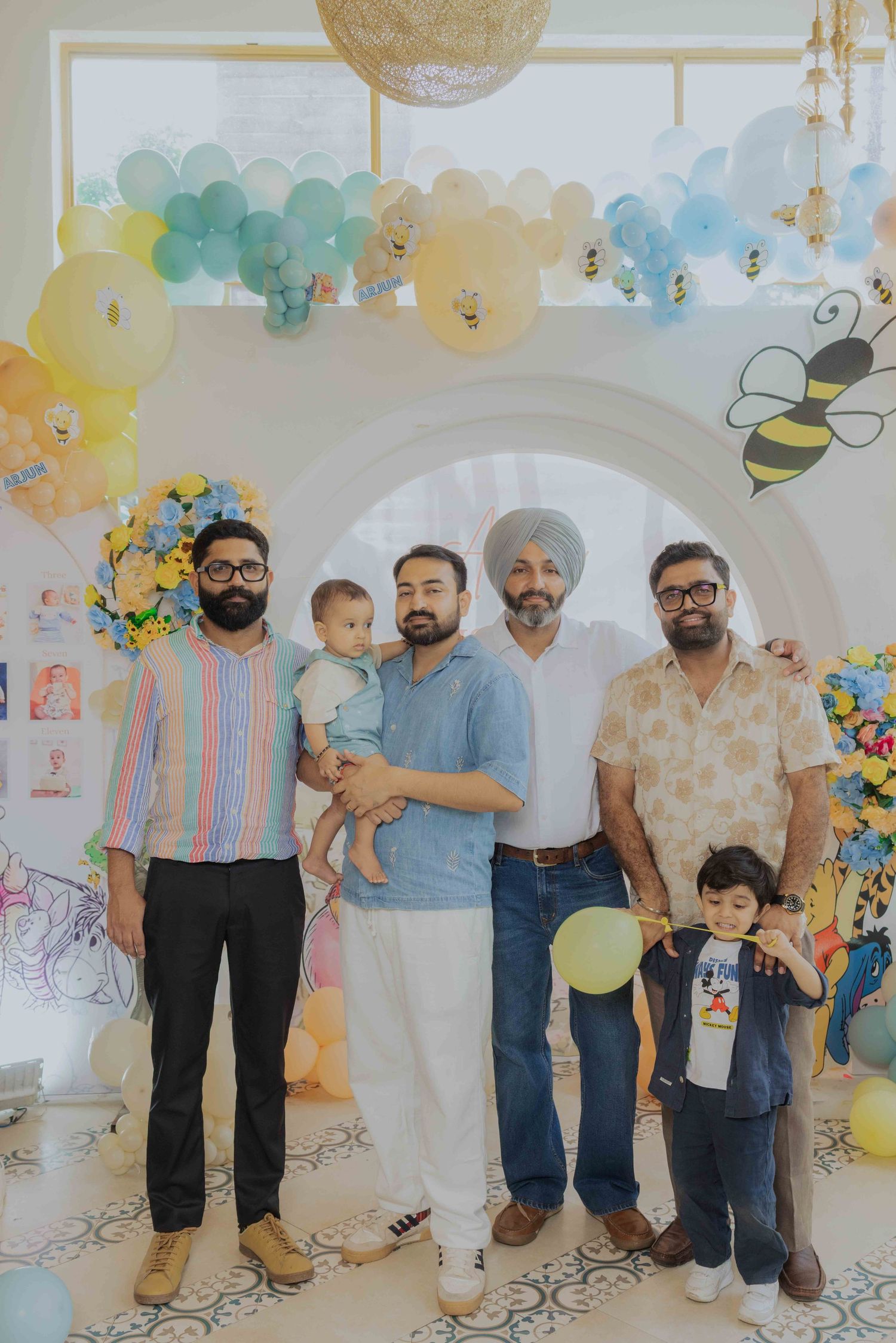 Four men and a toddler pose at a colorful bee-themed birthday party with balloon decorations.