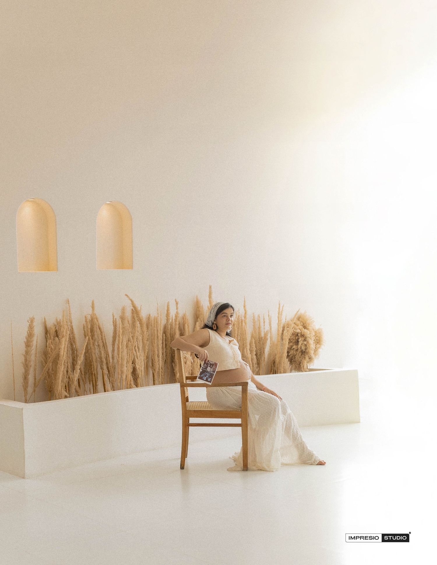 Pregnant woman in white dress sits on wooden chair surrounded by pampas grass in minimalist cream studio.