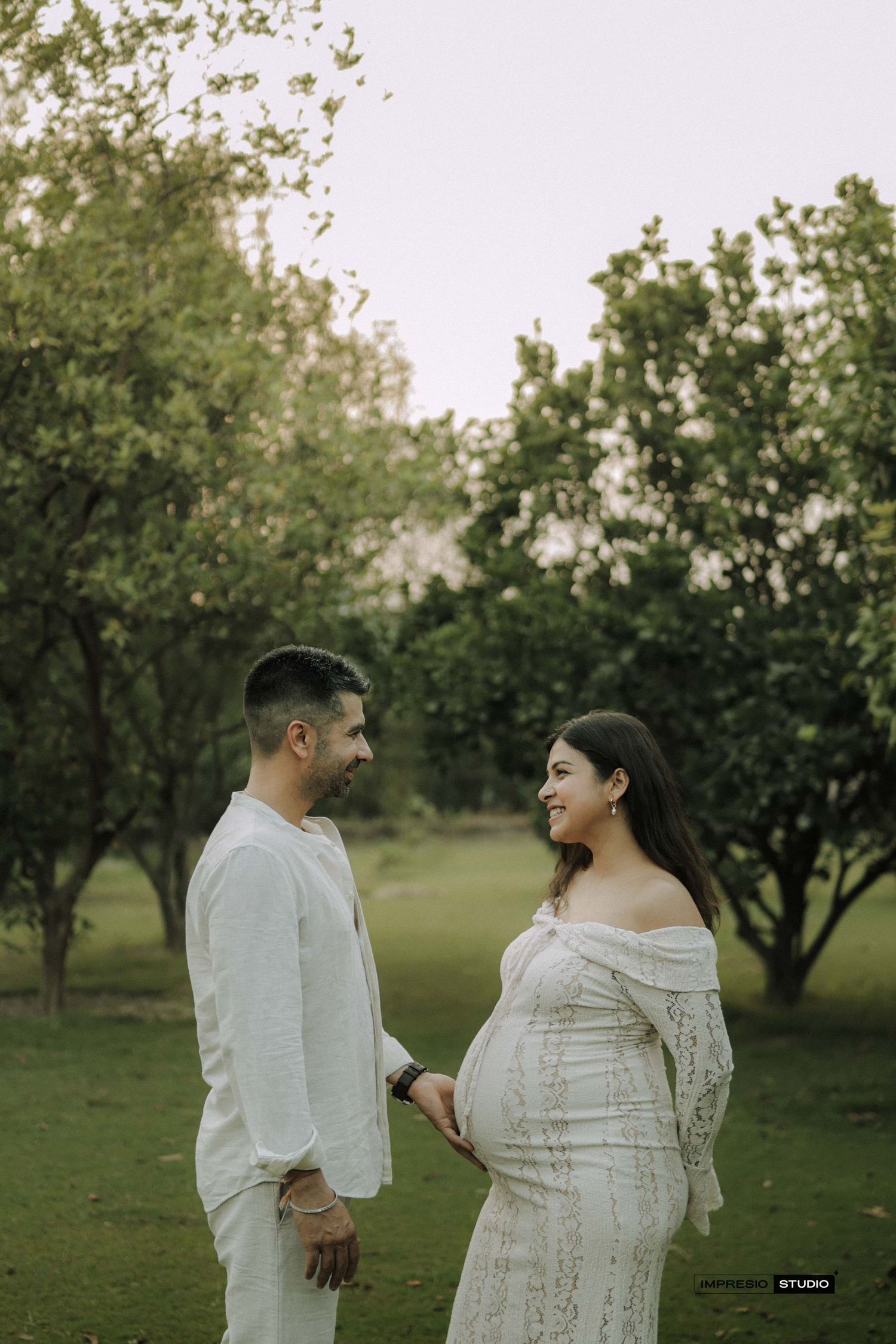 A couple in white outfits shares a tender moment outdoors, the woman cradling her baby bump in a lace maternity gown.