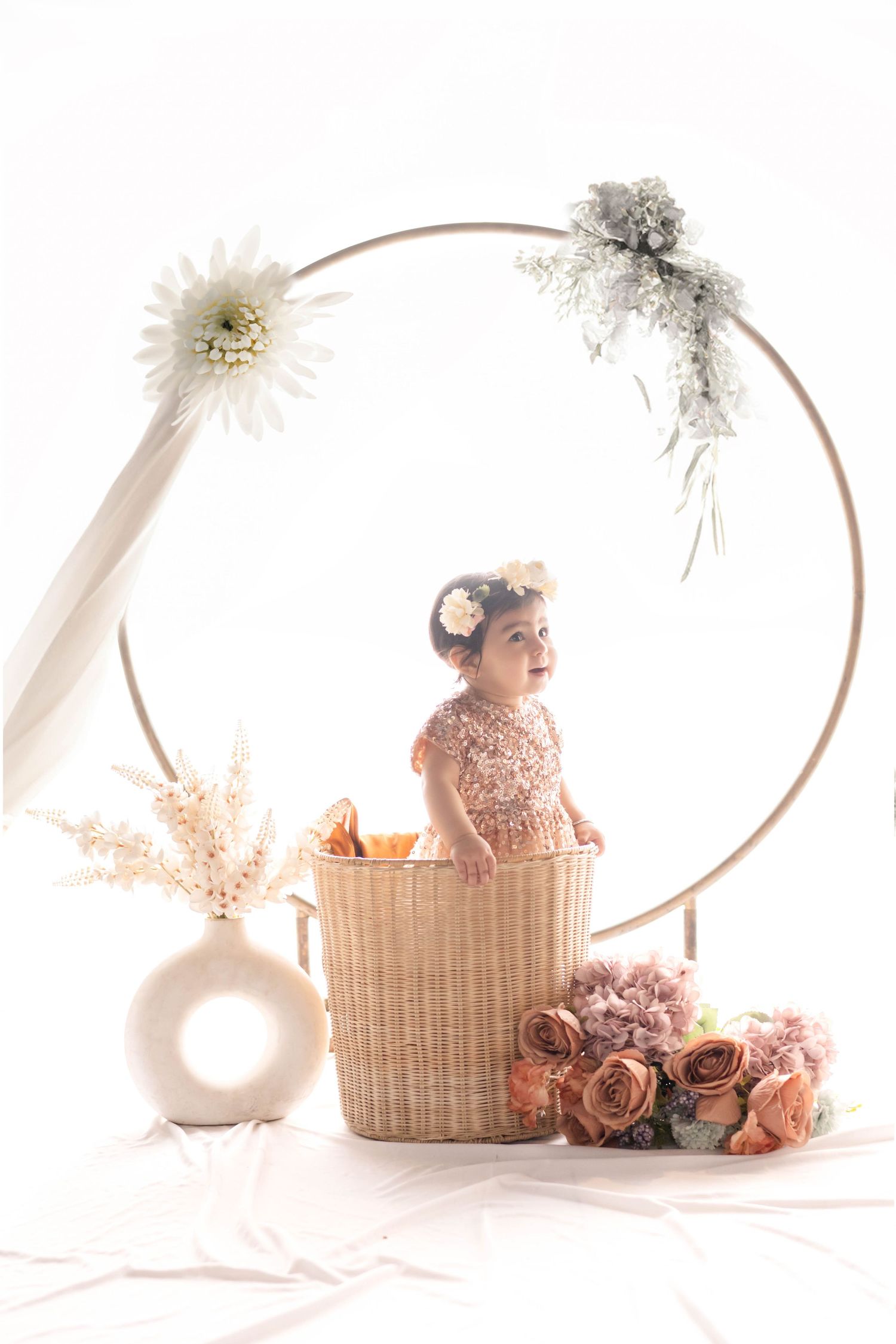 An adorable baby girl in a pink sequin outfit stands in a wicker basket surrounded by flowers and a floral hoop backdrop.
