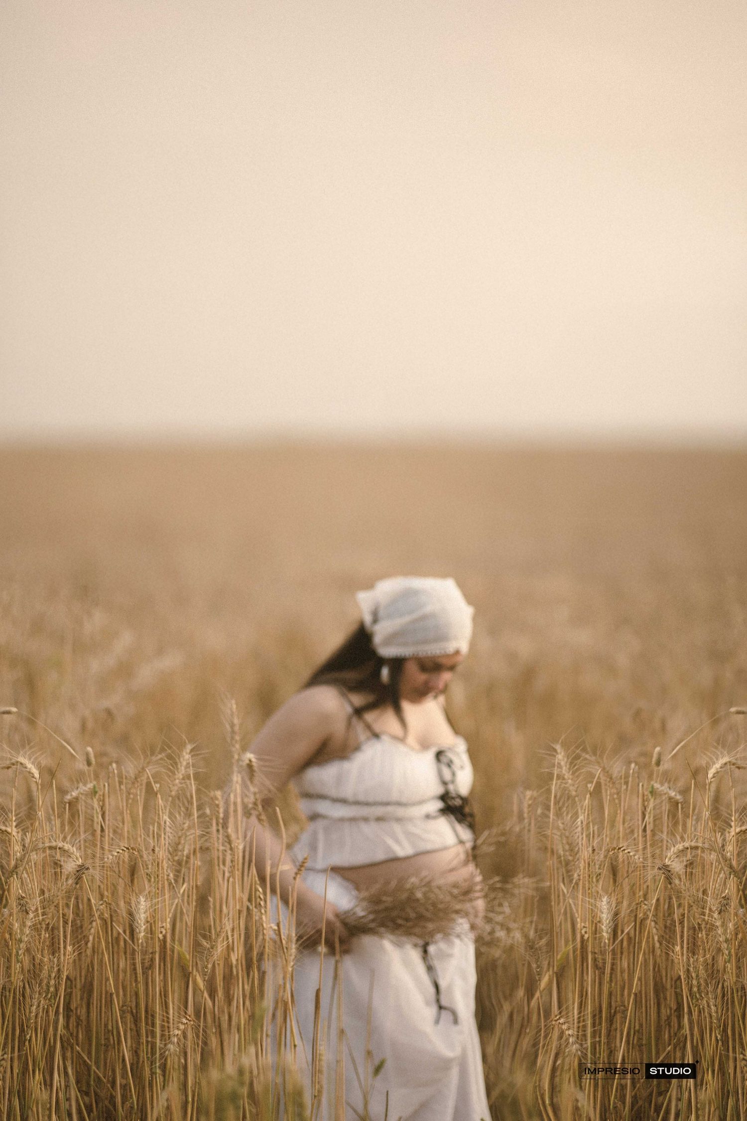 Pregnant woman in white dress standing in golden wheat field cradling her baby bump at sunset.