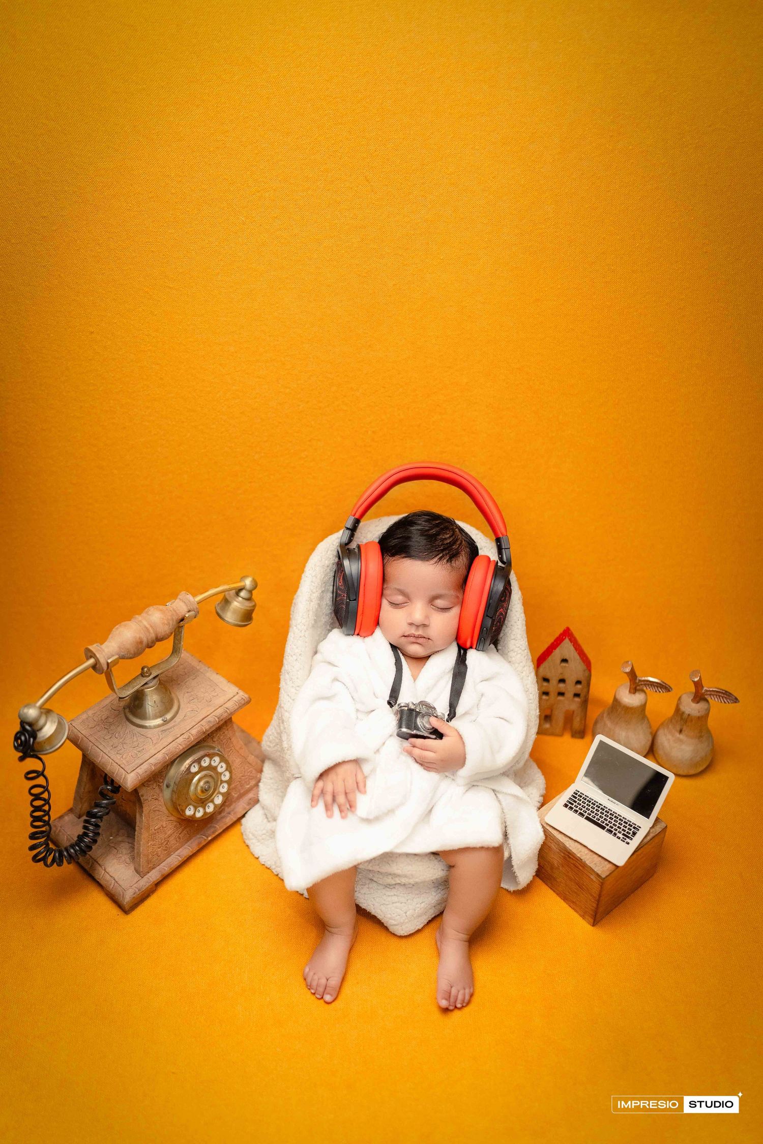 Baby in white robe wearing red headphones surrounded by vintage phone, laptop, and decor on yellow background.