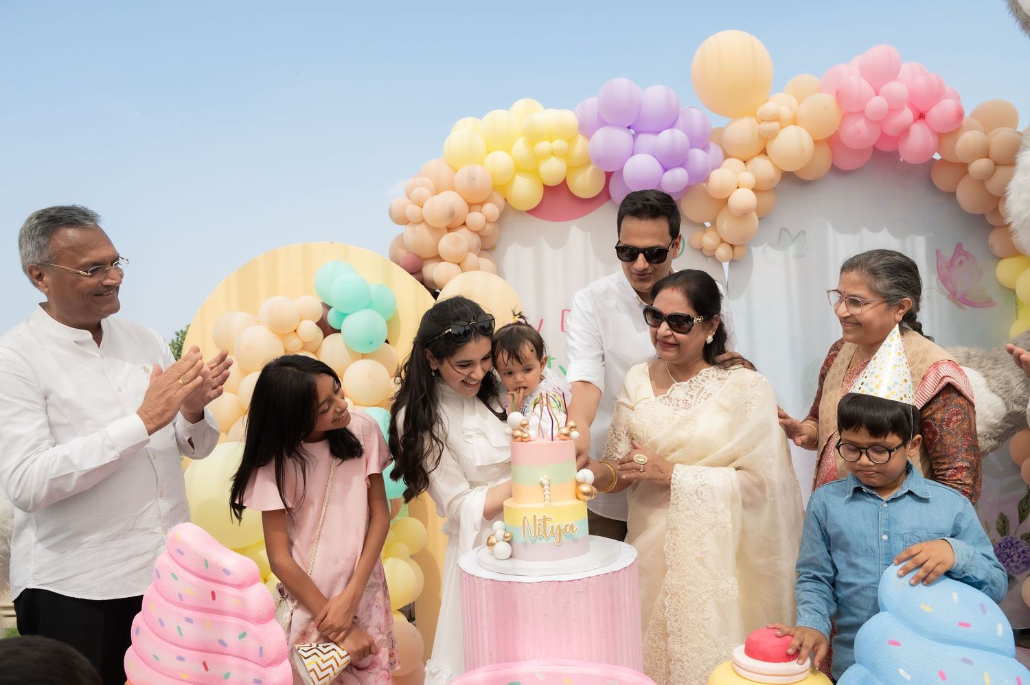 Family celebrating a baby's first birthday with a pastel balloon arch and cake outdoors.