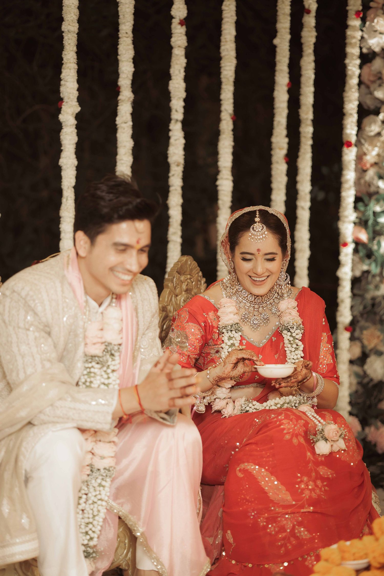 A smiling Indian bride in red lehenga and groom in cream sherwani sit together during traditional wedding ceremony.