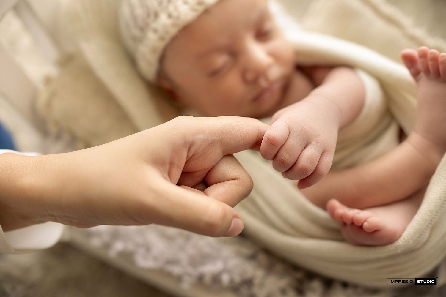 Newborn baby sleeping while gently gripping an adult's finger in a soft, wrapped newborn photography session.