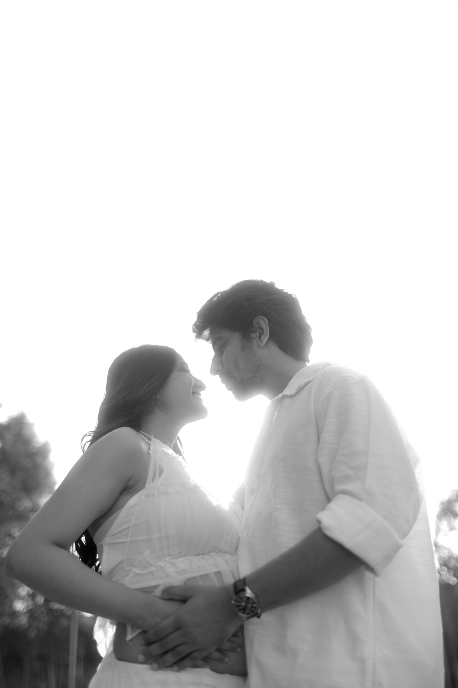 Black and white photo of a couple sharing a tender moment, with the man gently holding the womans pregnant belly outdoors.