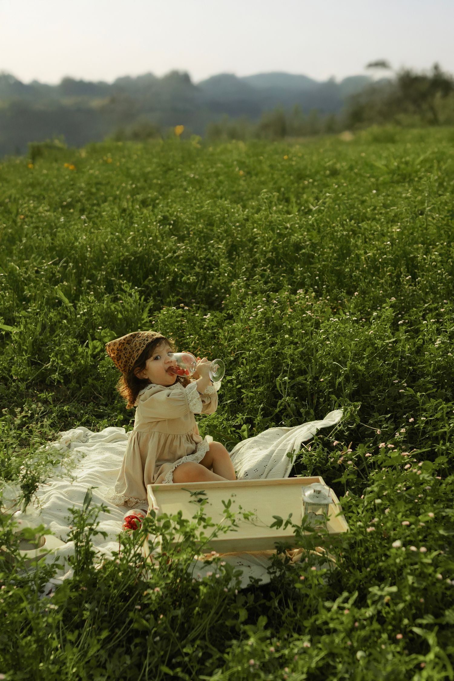 Toddler in straw hat and beige dress sitting in lush green meadow, drinking from bottle during outdoor picnic.