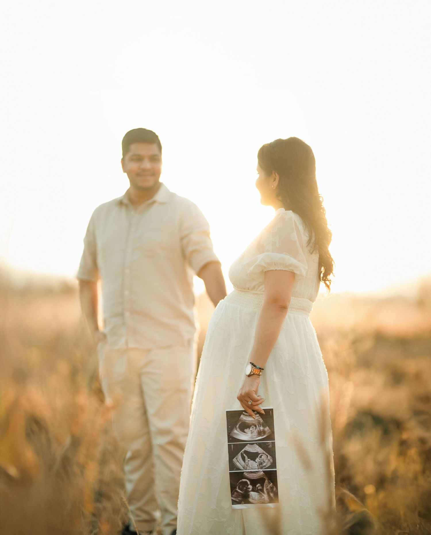 Expectant couple stands in golden sunlit field with ultrasound photos during maternity photoshoot at sunset.