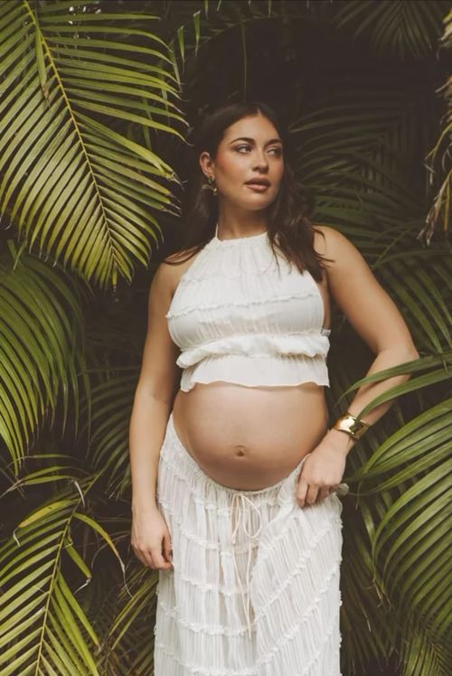 A pregnant woman in white lace dress poses against tropical palm fronds in a maternity photo session.