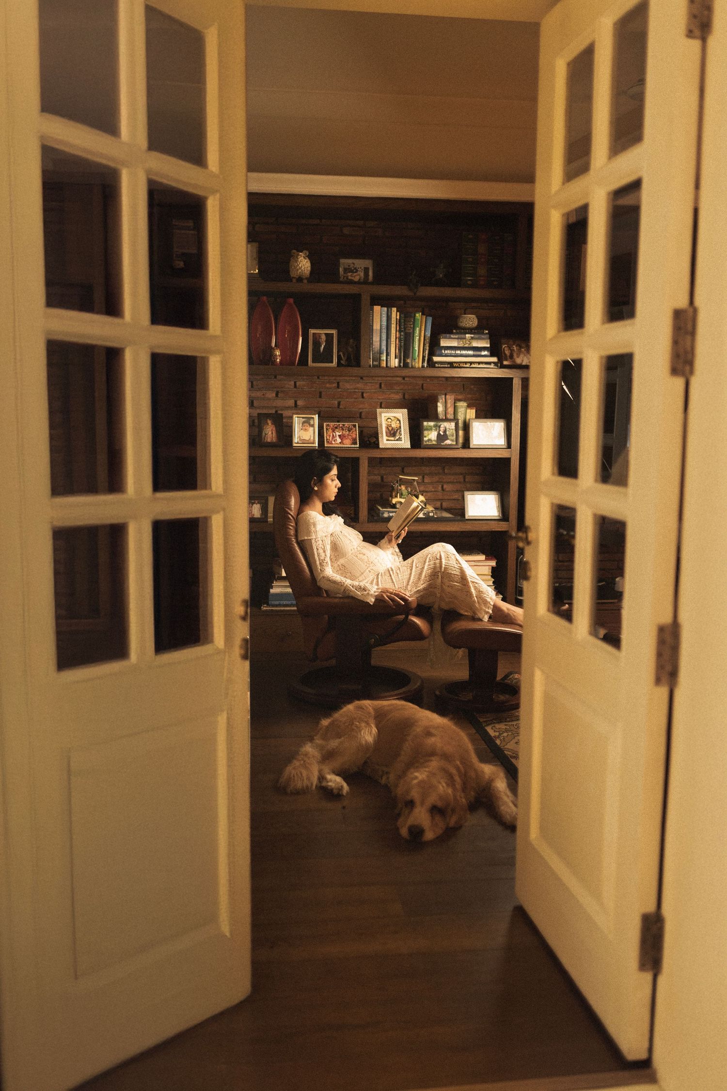 Through French doors, a cozy reading nook with bookshelves and a sleeping dog on dark hardwood floors in warm lighting.