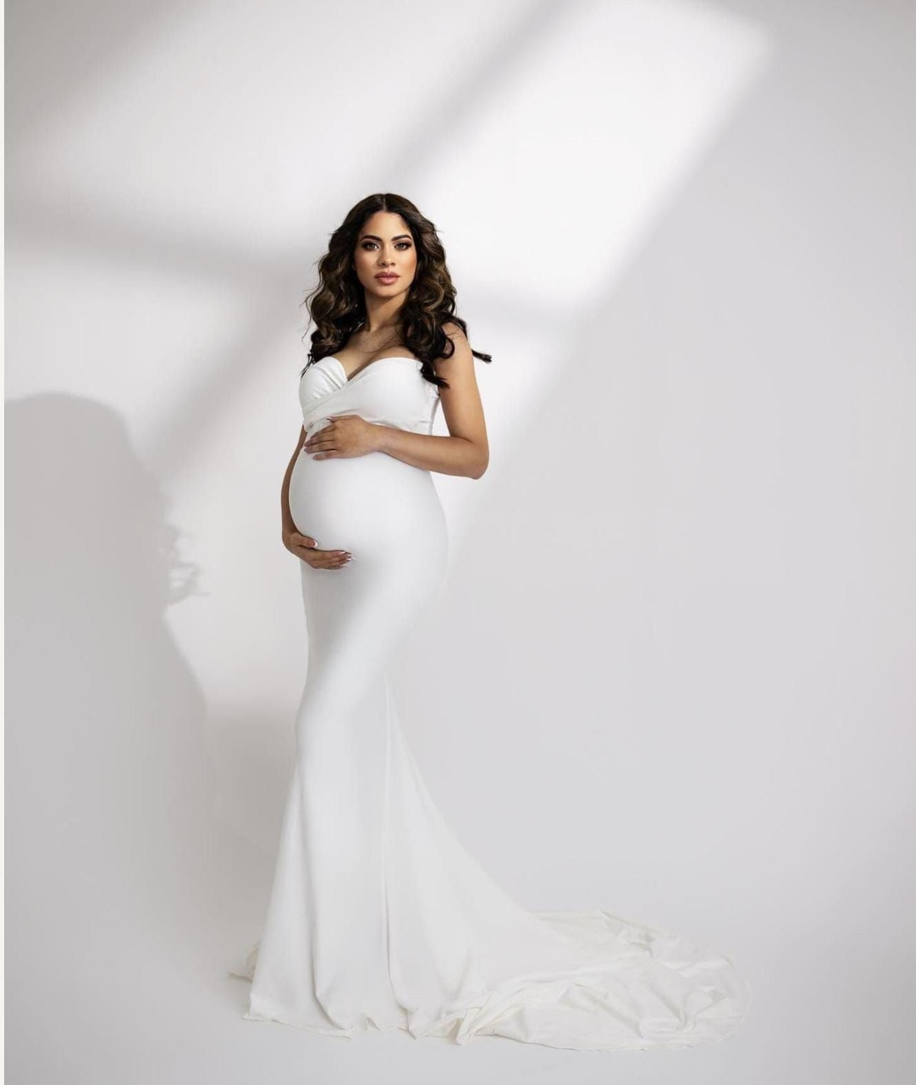 Minimalist maternity studio photo featuring white form-fitting gown against light backdrop.