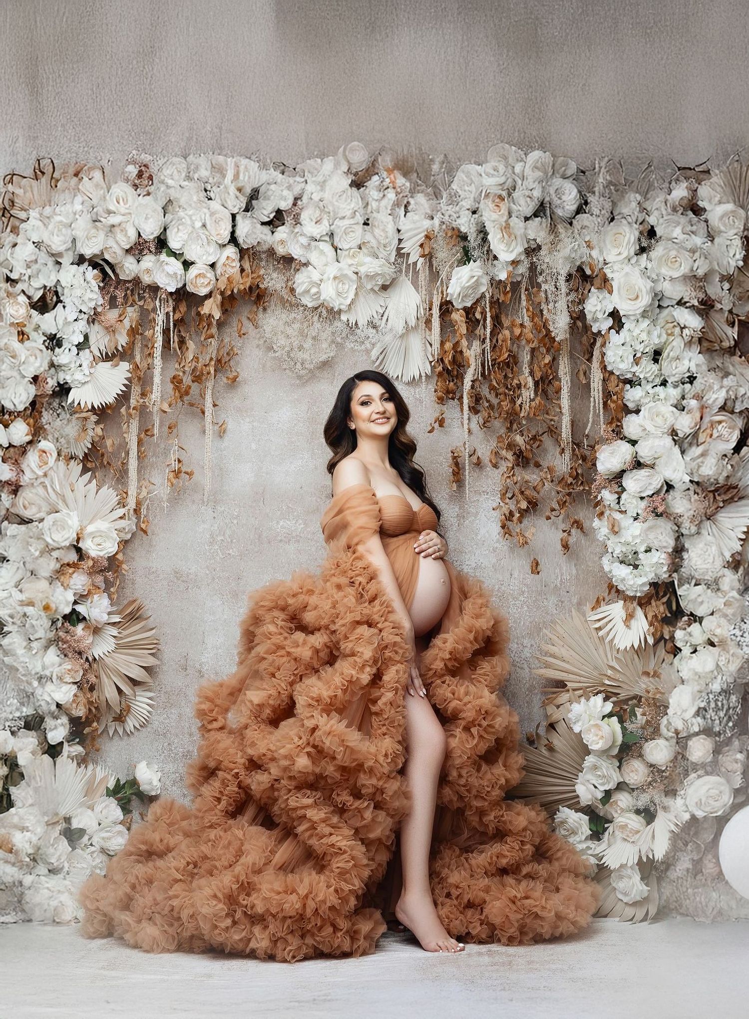 Dramatic maternity portrait with rust-colored tulle gown against white floral arch backdrop.