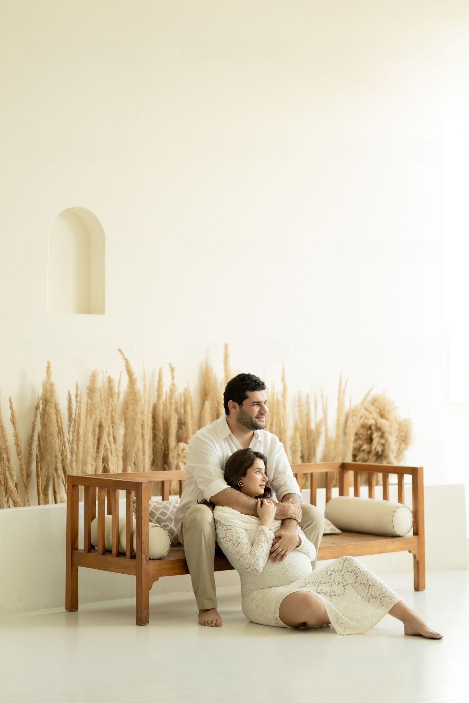 A minimalist white room features a person in casual attire reclining against a wall with wooden stakes arranged in a row.