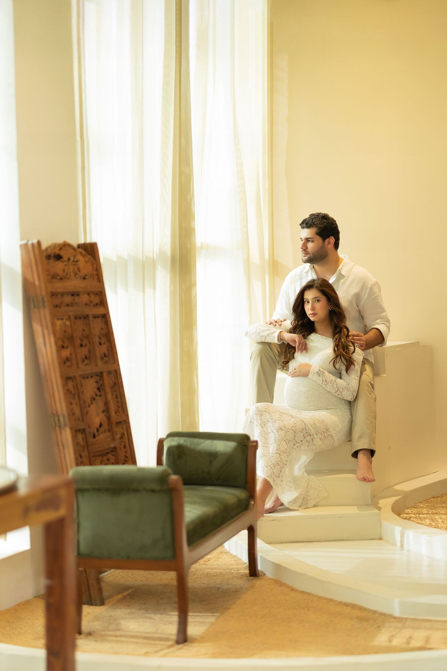 A couple sits together in a bright sunlit room with vintage green armchairs and white curtains.
