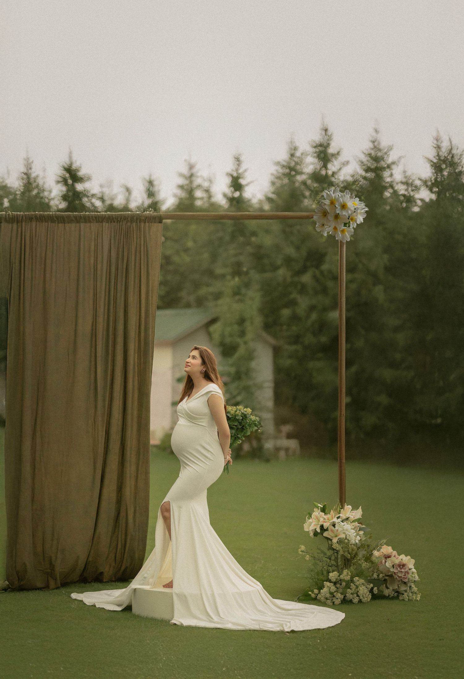 Elegant maternity photo with cream-colored flowing gown against forest backdrop and brown draping curtain outdoors.